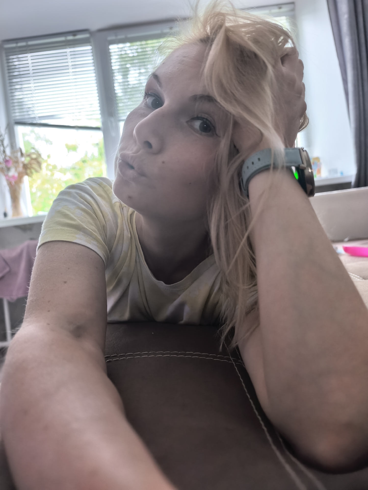 An informal, eye-level portrait shows a blonde woman posing playfully indoors. She has fair skin, light-colored eyes, and her blonde hair is casually styled, with some strands falling around her face. Her left hand is raised, gently touching her hair near her head, while she leans on her right arm, which is resting on a brown, possibly leather, surface with visible white stitching. She is wearing a light-colored, tie-dye t-shirt, possibly yellow or off-white. On her left wrist, she sports a smartwatch with a light blue band and a green light emitting from a sensor on its underside.
The woman is looking directly towards the camera with a "pouty" or "kissing" expression, suggesting a selfie or a casual photo. The natural light streaming in through the background windows indicates it is daytime, likely morning or afternoon. Behind her, a large window fitted with horizontal blinds offers a glimpse of lush green foliage outside, implying a season other than winter. To the far left, a vase containing dried flowers or foliage sits on a windowsill. Dark gray curtains are partially visible to the right of the window, framing the scene. Other indistinct objects, including some bottles on a surface and a pink item on a rack, can be seen further in the background. The overall setting appears to be a cozy domestic space, such as a living room, located in Sterlitamak, Russia.