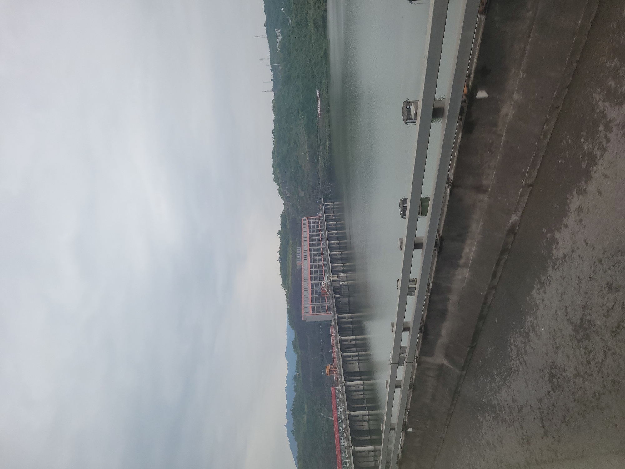 The image shows a gray body of water and a dam. The dam is on the right side of the image, made of concrete. There are metallic railings along the dam. In the distance, there is a red building structure extending into the water, with several parallel pipes beneath. The water reflects light and the sky. In the background on the left, there is a green forested hillside. The sky is gray and overcast. Caojie, China is the location.