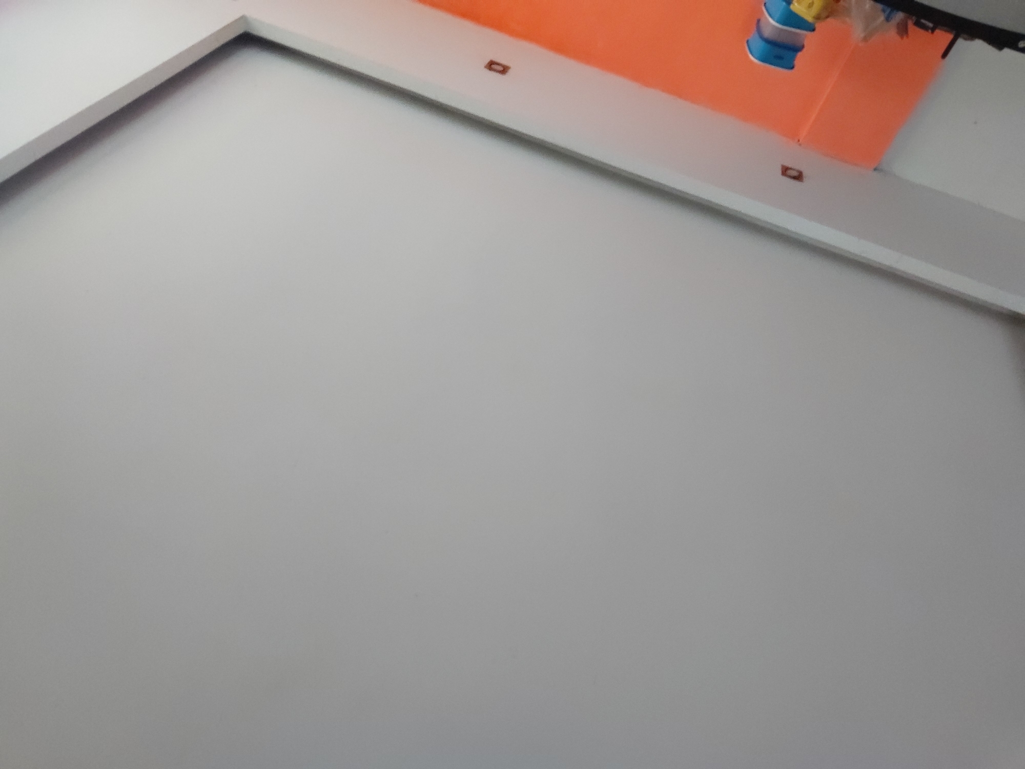 The image presents an interior scene, primarily featuring a large, plain, light gray or off-white wall that covers most of the frame. This wall appears to be a flat, unadorned surface.
At the top of the image, separating the main wall from an upper section, is a white horizontal trim or ceiling panel. Above this white trim, the upper wall or ceiling area is painted a vibrant orange.
Visible on the white trim are two small, dark, rectangular objects, possibly embedded spotlights or sensors, one towards the left and another further right. In the top right corner, hanging down from the orange section, is a blue and yellow object, which resembles a pet water dispenser or a similar container, partially covered in clear plastic packaging.
The photograph is taken from a low-angle perspective, looking upwards at the wall and ceiling. The lighting is uniform, suggesting an indoor environment without direct sunlight. There are no people visible, and no specific activity is taking place. No text is discernible in the image. The setting appears to be a functional room in Santa Ana, Venezuela.