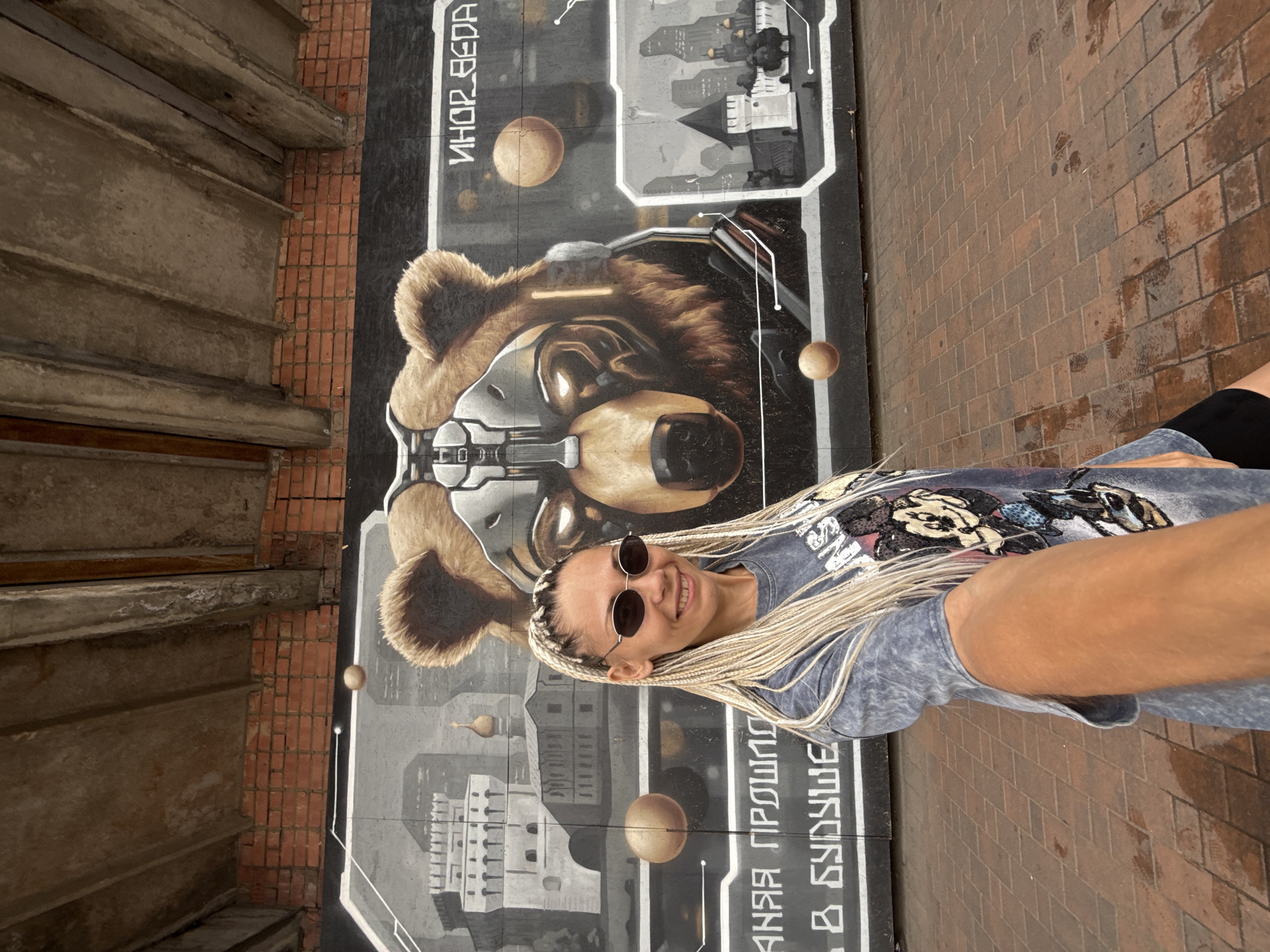 The image shows a young woman with blonde braided hair and sunglasses posing in front of a mural. She is wearing a gray t-shirt with a colorful graphic print and black shorts. The mural depicts a stylized bear's head with cybernetic elements and a futuristic city skyline in the background. There are also spheres scattered across the mural, possibly representing planets or other celestial bodies.
The woman is smiling and looking at the camera, suggesting a relaxed and happy mood. The setting appears to be outdoors, with brick pavement visible to the right of the mural and a concrete structure with horizontal beams on the left. The lighting suggests it is daytime, possibly late morning or early afternoon, and the weather seems clear.
Visible text on the mural includes Cyrillic script, with the words "ИНОР_ВЕДА" at the top and "АНАЯ ПРОШЛО" and "В БУДУЩЕ" near the bottom. The overall impression is of a vibrant urban art installation with a young woman enjoying the artwork. The location context of Rusanovo, Russia, is noted.