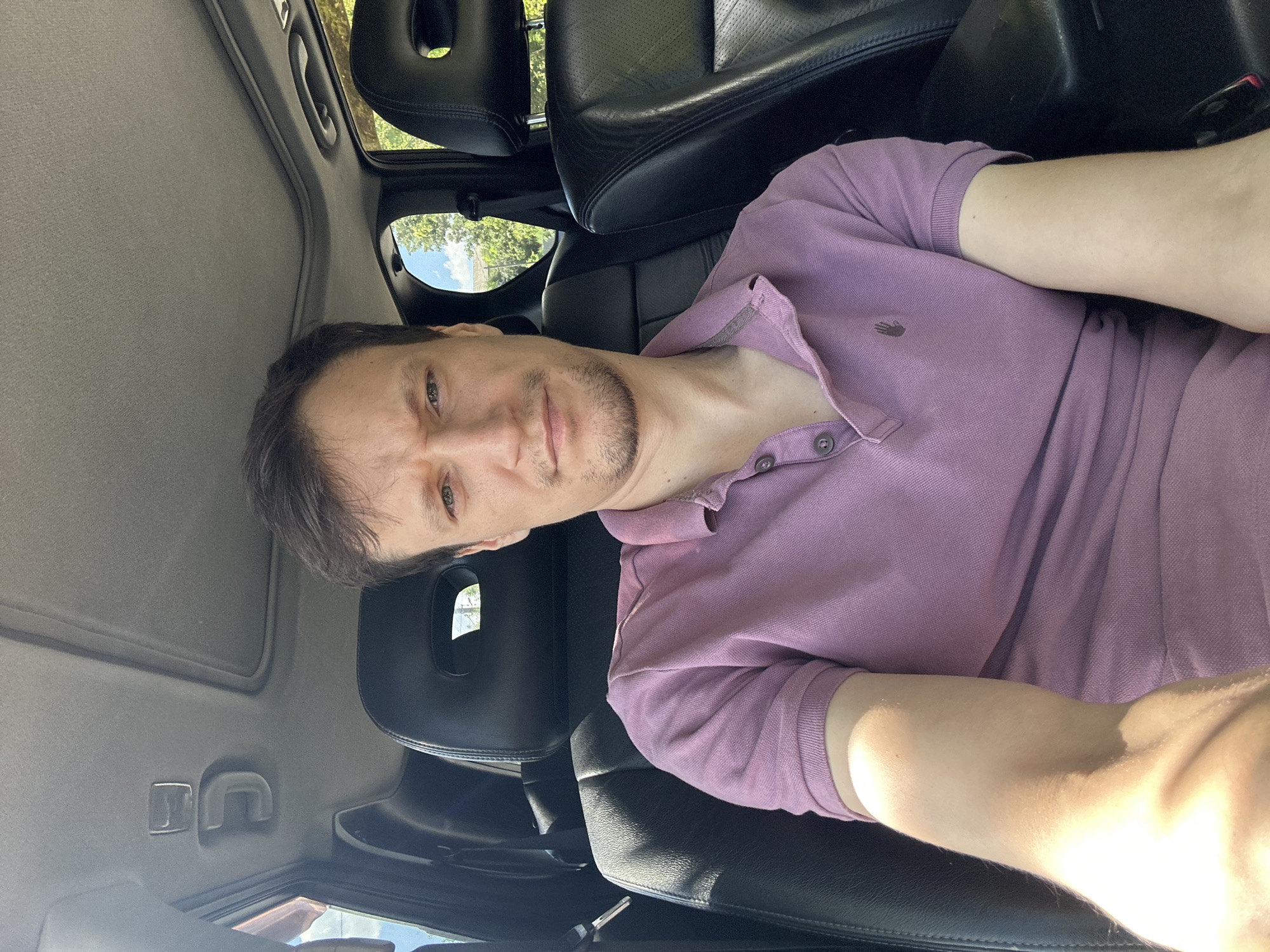 A man is sitting in the driver's seat of a car, facing forward and looking at the camera. He is wearing a purple polo shirt. The car's interior, including black leather seats and a sunroof, is visible. Outside the car, trees and a blue sky can be seen. The scene is outdoors during the daytime. The provided information about the location of Makiv, Ukraine, cannot be verified from the image alone.