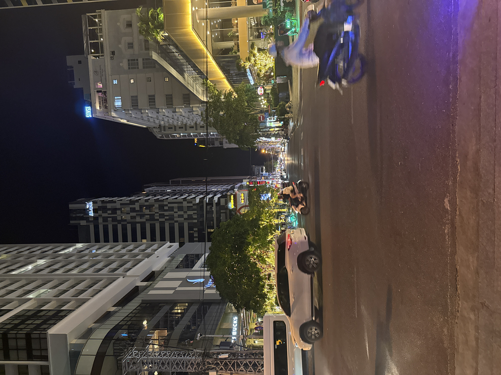 The image depicts a street scene in Nha Trang Ward, Vietnam, at night. The scene is vertically oriented. High-rise buildings are visible on both sides of the street, illuminated by streetlights and building lights. Street-level businesses, including one with "BUCKS" visible in its storefront sign, are also lit. Several vehicles are present on the road, including a white car parked on the right side of the frame. Motorbikes with riders are moving along the street. The road surface shows signs of wear and potential rain. The sky is dark, indicating nighttime. Overall, it's a common urban nighttime scene.