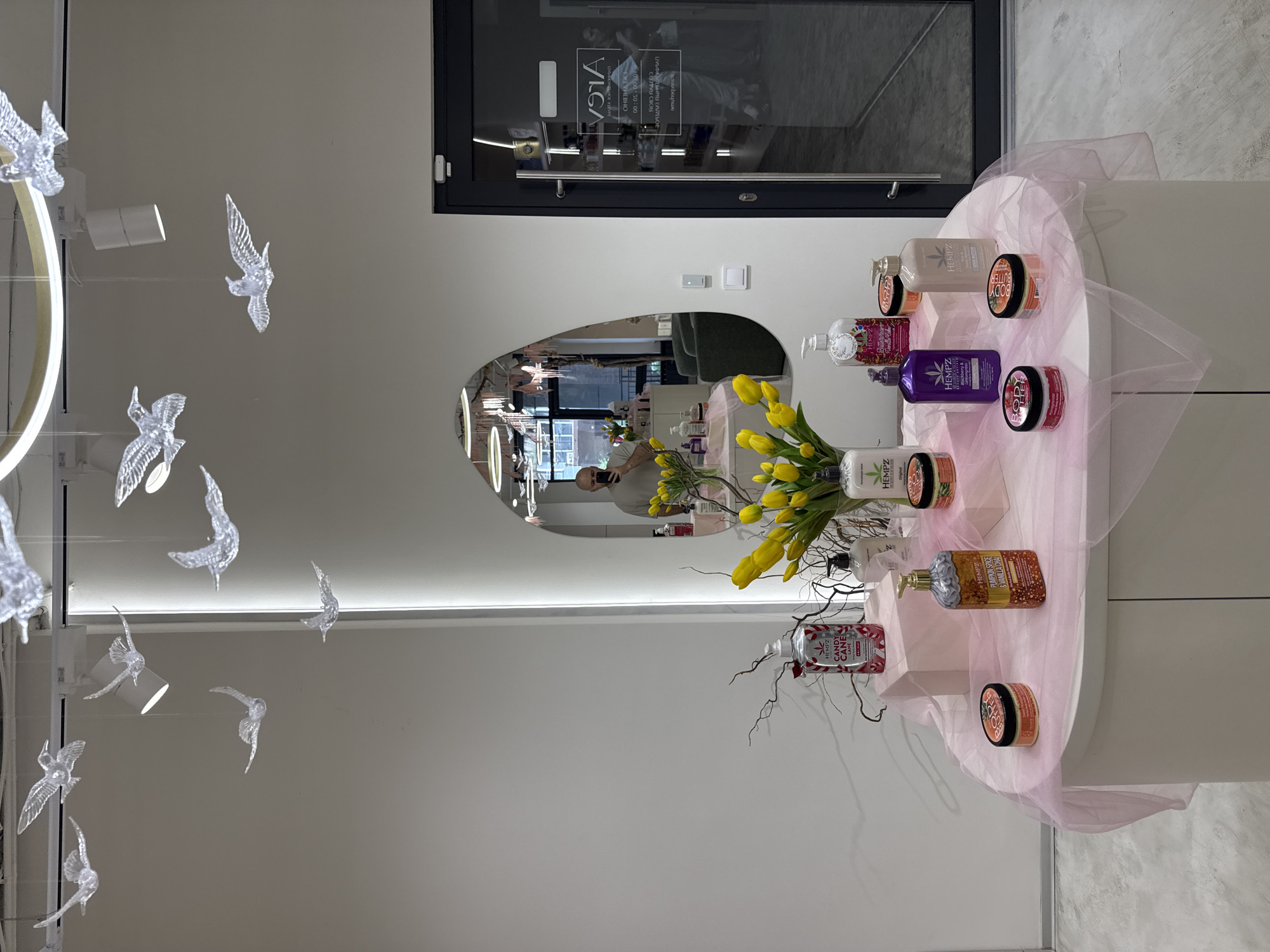 An indoor retail or salon space is depicted with a focus on a display table in the foreground. The table is round and white, covered with a sheer pink fabric. Arranged on the table are various products, primarily bottles and jars of lotions and body butters, many featuring the "HEMPZ" brand. Among the products, a vase of bright yellow tulips serves as a central decorative element. Behind the display table, a large, irregularly shaped mirror reflects the interior of the space, including a man taking a photograph with his phone. The background shows a clean, modern aesthetic with white walls, and an illuminated pillar. To the right, a glass-paneled door with black framing leads to another area, with signage visible on the glass indicating "Arca" and operating hours. Decorative, clear bird-shaped ornaments are suspended from the ceiling by thin wires, adding an artistic touch. The overall impression is of a well-organized and aesthetically pleasing retail environment.
