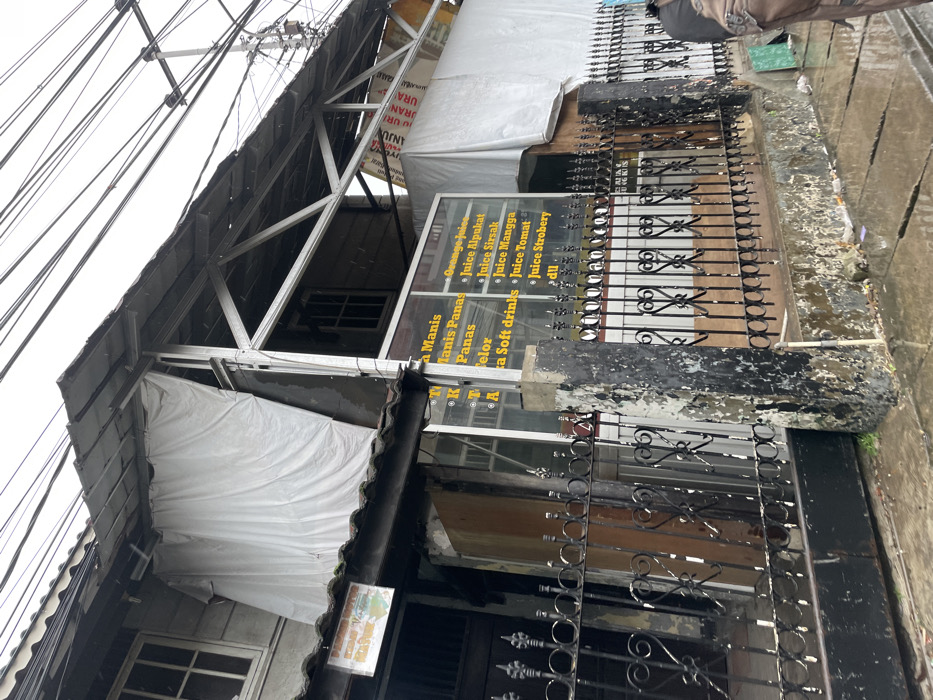 The image shows a storefront in Ngamprah, Indonesia (latitude: -6.863, longitude: 107.502). The storefront is made of black metal and has a white awning above it. The awning is supported by a metal frame. The storefront has a sign that lists various beverages for sale in Indonesian. The storefront is closed, and there is a black metal fence in front of it. The ground in front of the storefront is wet, and there are puddles of water. A person is walking on the sidewalk behind the fence, but their face is obscured. The sky is overcast. The image was taken from a low angle, and the camera is pointing upwards.