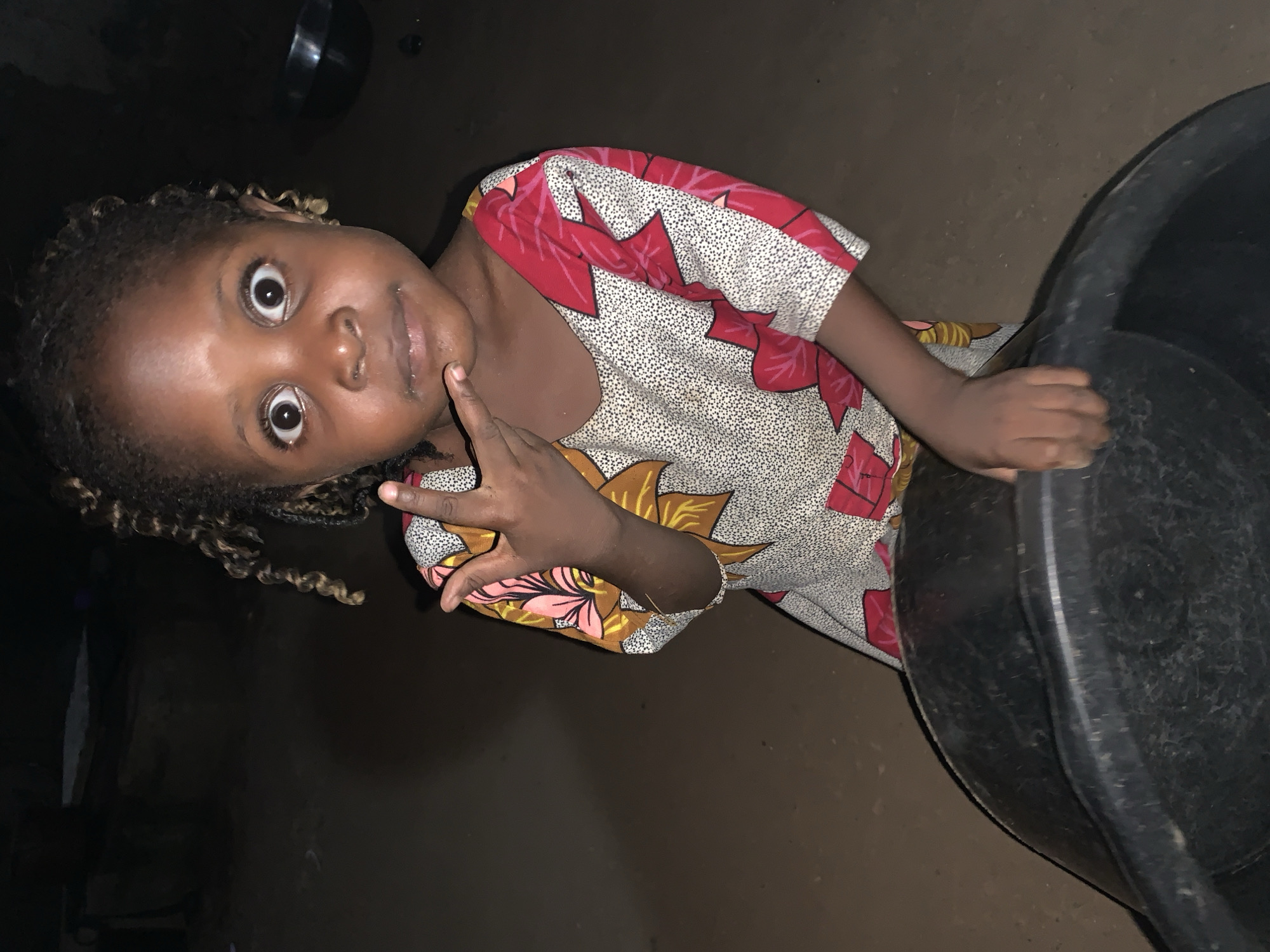 A young girl with curly hair stands indoors in Agban, Nigeria, wearing a patterned dress. She holds up her left hand, making a gesture with her fingers. A black tub is to her right. The lighting is dim.