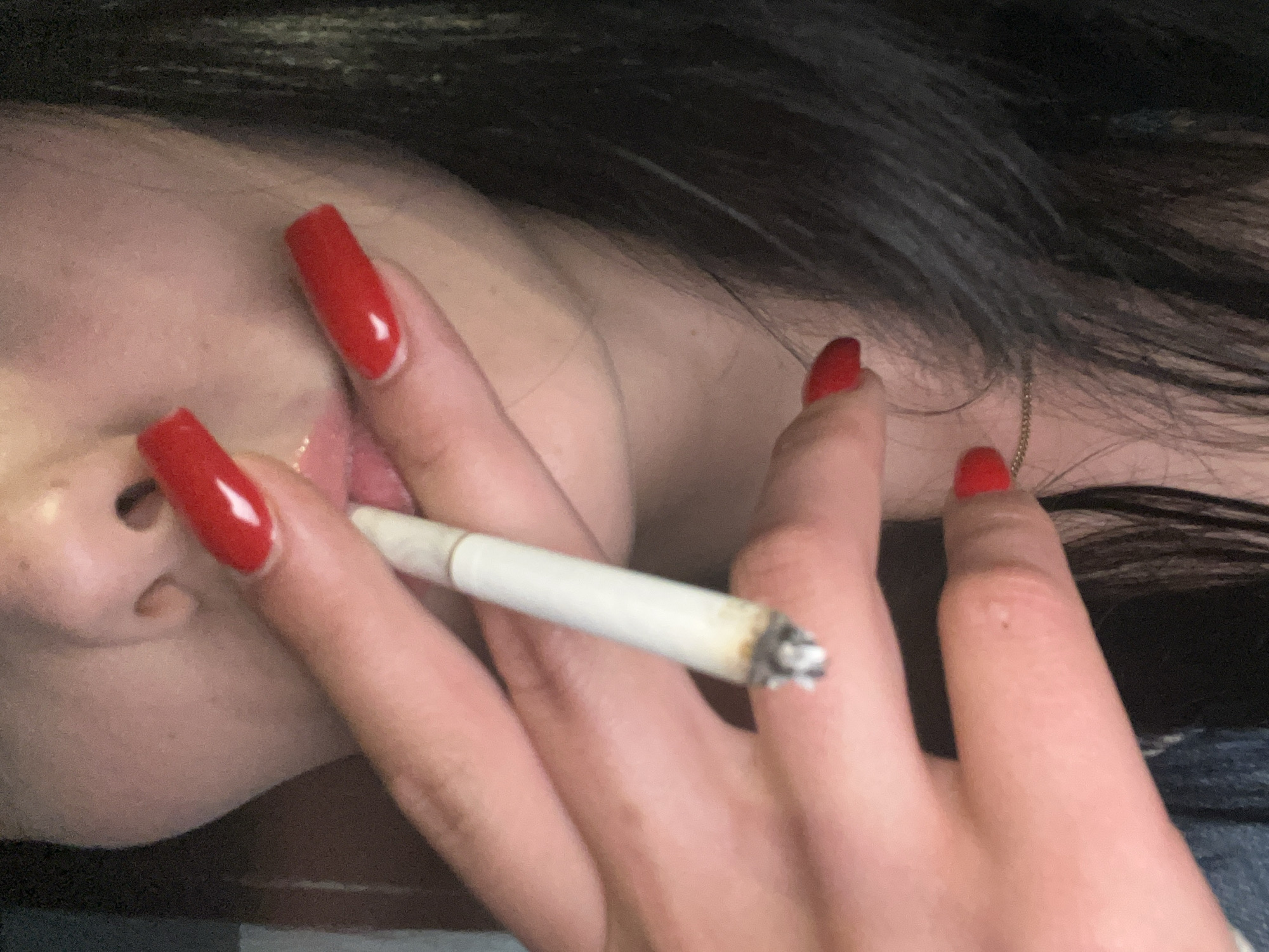 A close-up, angled shot captures a person holding a lit cigarette. The lower part of the person's face is visible on the left, showing the nose, partially open mouth, and chin area, with light skin tone. Their hand, adorned with long, bright red, square-tipped fingernails, holds the cigarette between the index and middle fingers. A faint, lighter-colored detail is noticeable on the base of one red nail, possibly a French tip.
The cigarette is partially smoked, displaying a visible ash tip and a clean filter end positioned near the person's lips. Dark, seemingly long hair is visible in the background, flowing around the neck and shoulder area, partially obscuring a thin gold chain necklace worn around the neck. The scene appears to be dimly lit, creating a soft and somewhat intimate atmosphere. There is no visible text in the image.
