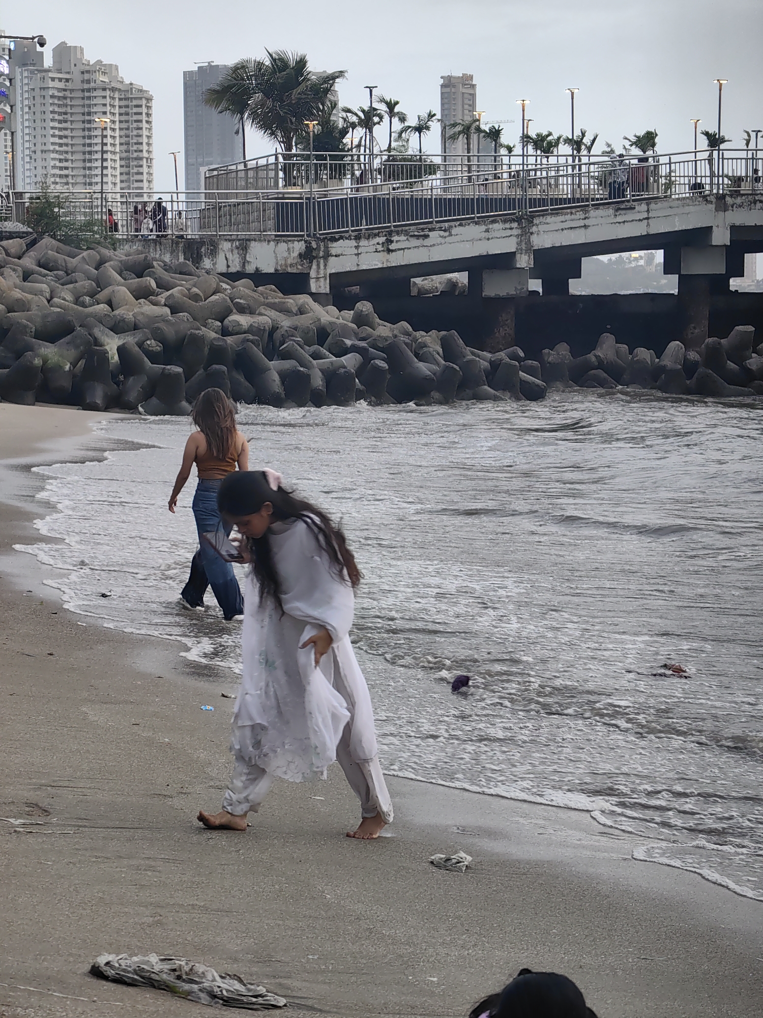 Two young women are walking on a beach in Mumbai, India, near a large, concrete breakwater and a pedestrian bridge. The woman in the foreground is wearing a white shalwar kameez and walking barefoot towards the left of the frame, looking down at her phone. The woman in the background is wearing a brown crop top and blue jeans and is walking barefoot towards the right of the frame, away from the camera. In the background, several tall buildings and palm trees are visible. The water is choppy, and there is some trash on the beach and in the water.