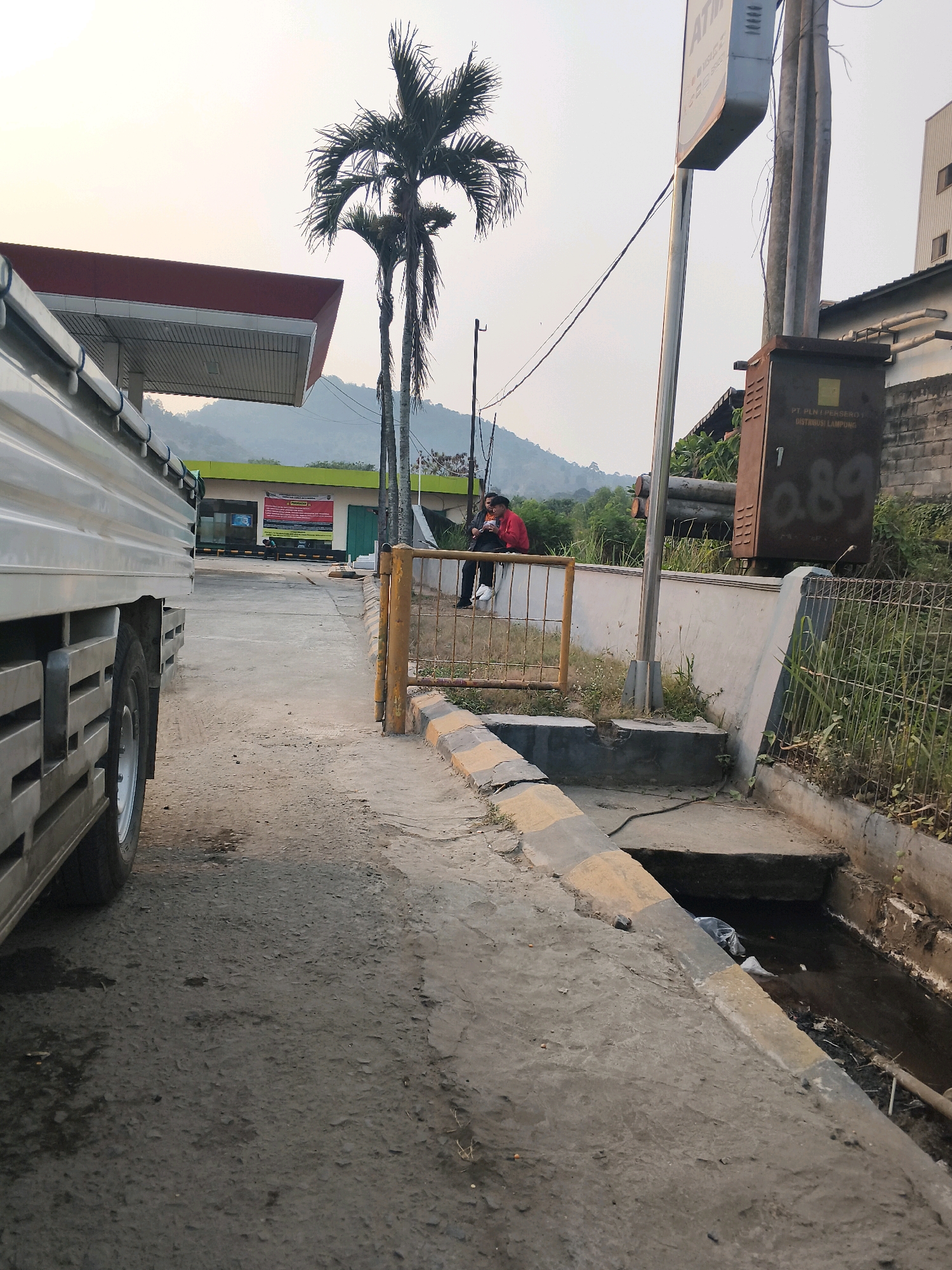The image shows a street scene in Sabah Balau, Indonesia. A white truck is parked on the left side of the frame, partially obscuring a gas station with a red roof. Two individuals are seated on a low wall in the background, one wearing a red jacket and the other a dark shirt. Behind them, two palm trees stand tall. A metal fence separates the walkway from a drainage ditch filled with murky water and debris. A large electrical transformer box is mounted on a pole to the right, with the label "PT PLN (PERSERO) DISTRIBUSI LAMPUNG" visible. The overall environment appears to be a semi-urban area with some vegetation and a hazy sky.