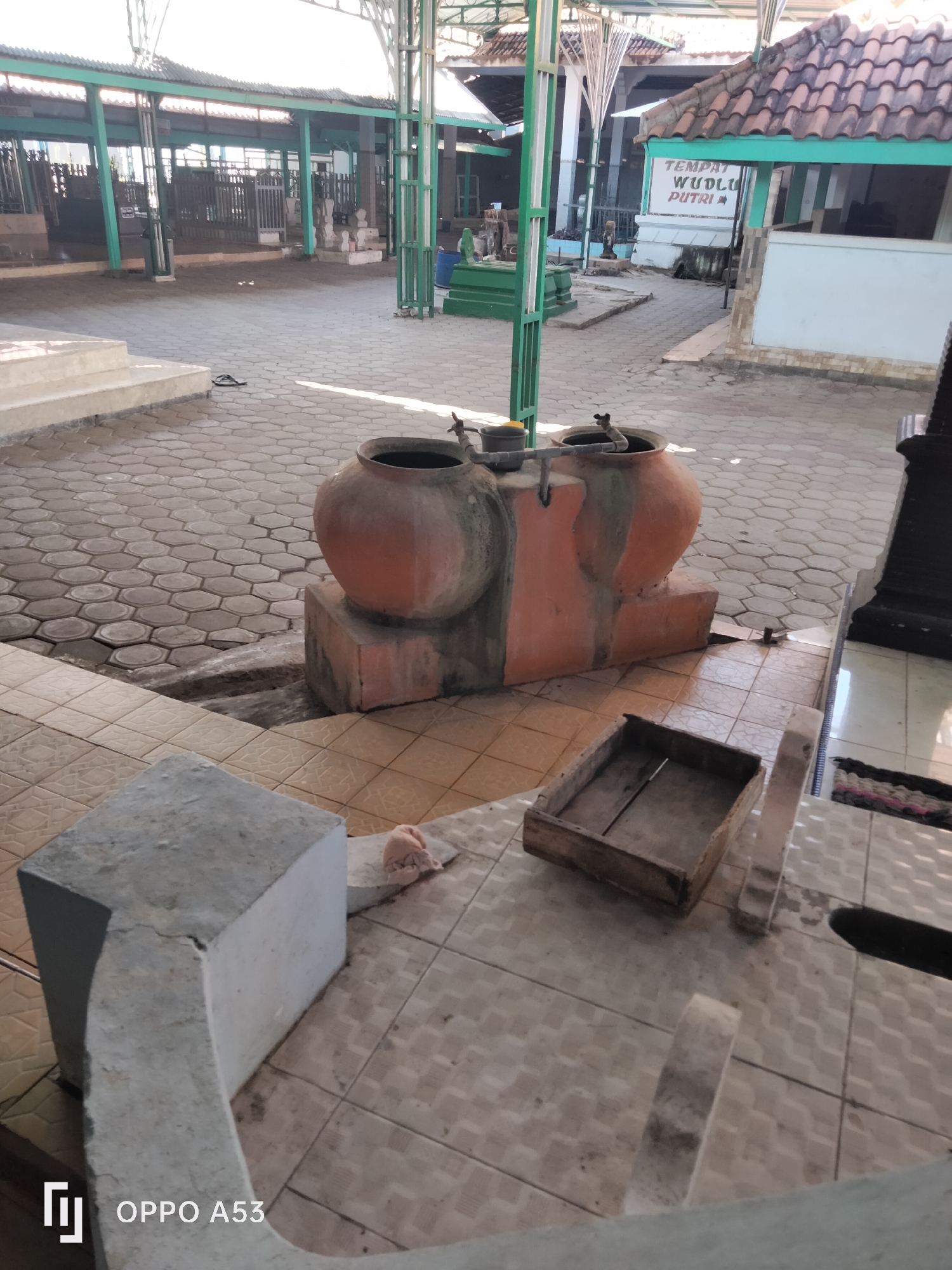 The image shows an outdoor area with a paved ground made of hexagonal and square tiles. In the center, there are two large, terracotta-colored clay pots with spigots, mounted on a concrete base. A wooden crate is placed on the tiled ground in front of the pots. In the background, a building with a tiled roof and a green roof overhang is visible, along with a sign that reads "TEMPAT WUDLU PUTRI". A concrete structure with stairs is visible on the left side of the image, and a flip-flop is lying on the ground near the stairs. The overall environment appears to be a public space, possibly a religious site or a place for ritual ablution. The image does not show any people, actions, or interactions.