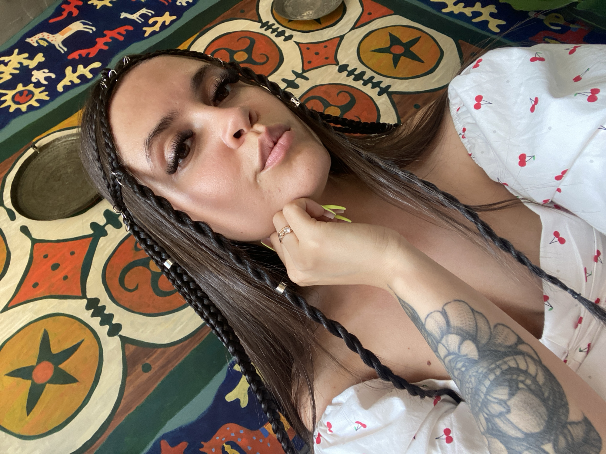 A close-up, high-angle selfie shows a young woman with dark hair styled in multiple braids with small silver rings woven in. She is wearing a white off-the-shoulder top with a red cherry pattern. The woman has her left hand on her chin and is wearing a ring on her left ring finger. Her fingernails are painted bright yellow. She has long, dark eyelashes and is looking directly at the camera. The background is a colorful, patterned tapestry or rug featuring geometric shapes, stars, and animal motifs. Based on the distinctive artistic style of the background, this image was likely taken in the city of Makhachkala, Russia, as the patterns are reminiscent of traditional Dagestani textile art.