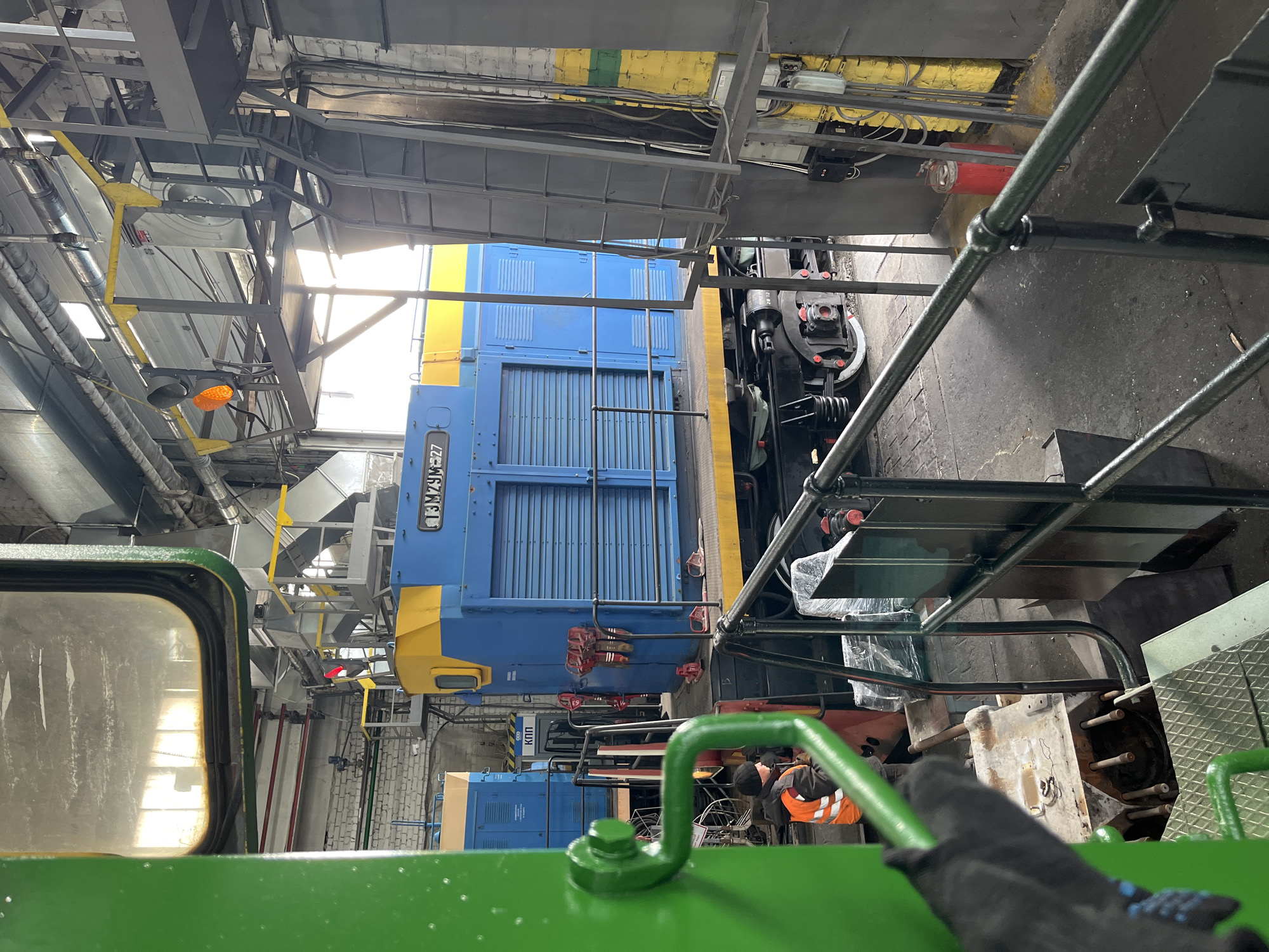 The image depicts the interior of a train repair facility in Chaikovsky, Russia. The primary subjects are two blue and yellow diesel locomotives, likely under maintenance. A person wearing a high-visibility orange vest is visible near the lower locomotive, possibly engaged in repair work. The environment is industrial, with overhead scaffolding, various pipes and wiring, and steel walkways. Green safety rails are prominent in the foreground, suggesting the viewpoint is from an elevated position within the facility. The overall impression is one of a working rail maintenance depot.