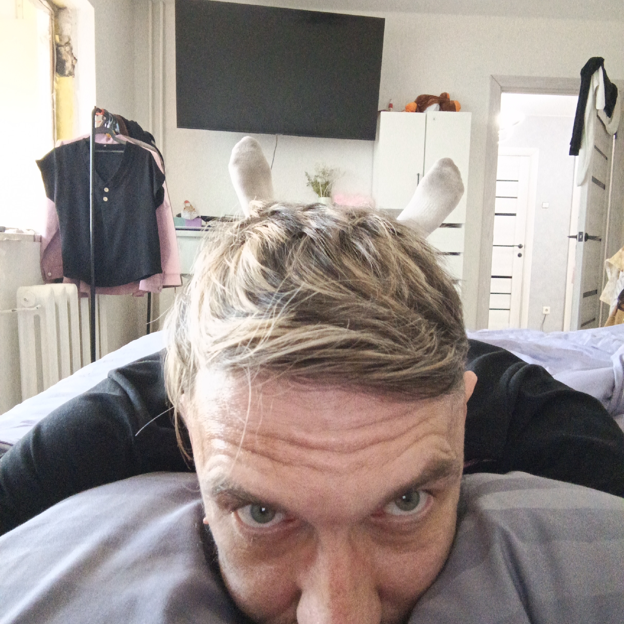 A close-up selfie taken by a man with short, light brown and blonde hair, grey eyes, and fair skin. Two white socks stand upright on his head, resembling bunny ears. He is lying on a bed with grey bedding. Behind him is a room with a black television mounted on the wall, a white cabinet, a radiator, and a doorway leading to another room. Clothes are hanging on a rack near the radiator. The location is likely residential, potentially in Марковский, Russia.