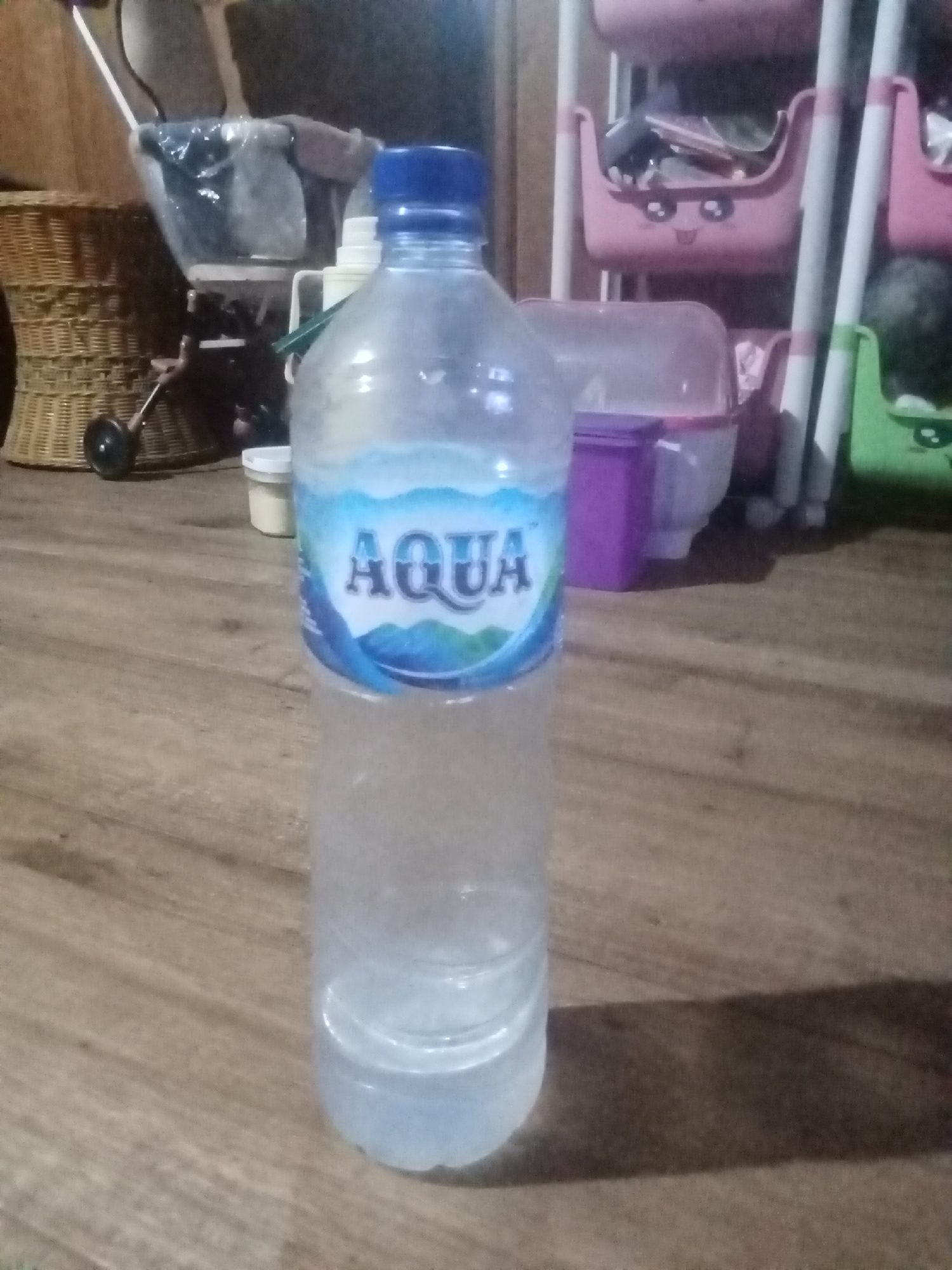 In a dimly lit indoor setting, possibly a residential area in Setia Jaya, Indonesia, a clear plastic bottle of AQUA brand bottled water stands upright on a wooden floor. The bottle is partially filled with water and features a blue cap and a label with the AQUA logo. In the background, a woven basket is visible to the left, and a multi-tiered plastic storage unit with pink and green compartments is to the right. Other objects, including what appears to be a stroller or baby carrier and various containers, are scattered throughout the scene, suggesting a cluttered living space.