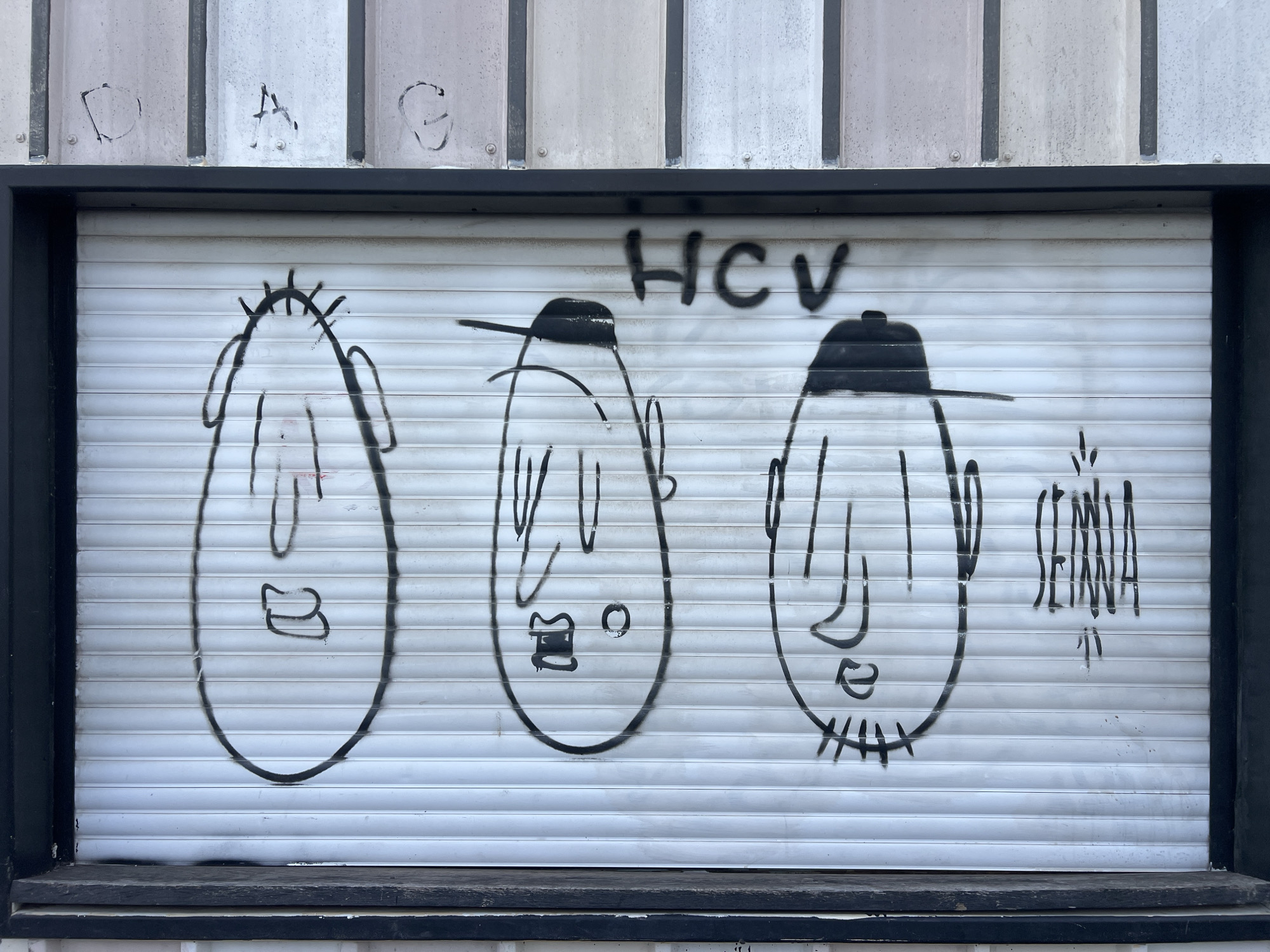 The image displays a white, horizontal corrugated metal roll-down security shutter, centrally framed by a dark, likely black, border, characteristic of a commercial storefront in an urban setting within Tel-Aviv, Israel. The shutter is adorned with black spray-paint graffiti featuring three stylized, anthropomorphic head-like figures arranged horizontally.
The leftmost figure is an elongated oval shape with spiky projections on top, simple line-segment eyes, a curved nose, and a small U-shaped mouth. The middle figure is a slightly narrower oval, topped with a solid black, flat-brimmed cap, two vertical lines for eyes, a small rectangular nose, and a small circular mouth. The rightmost figure is a similar oval, also wearing a solid black, flat-brimmed cap, with two vertical lines for eyes, a curved nose, and multiple short horizontal lines beneath the mouth, suggesting facial hair.
Above the middle and right figures, the uppercase letters "HCV" are spray-painted in black. To the immediate right of the rightmost figure, a vertical, abstract tag, possibly reading "SENNA" or "SEMNA", is visible.
Above the dark frame of the shutter, the building's facade consists of light-colored, vertically-oriented corrugated metal panels secured with visible rivets. Faint, stylized black graffiti letters, appearing as "D," "A," and "C" or "G," are present on these upper panels from left to right. The bottom edge of the shutter rests on a dark, horizontal ledge.