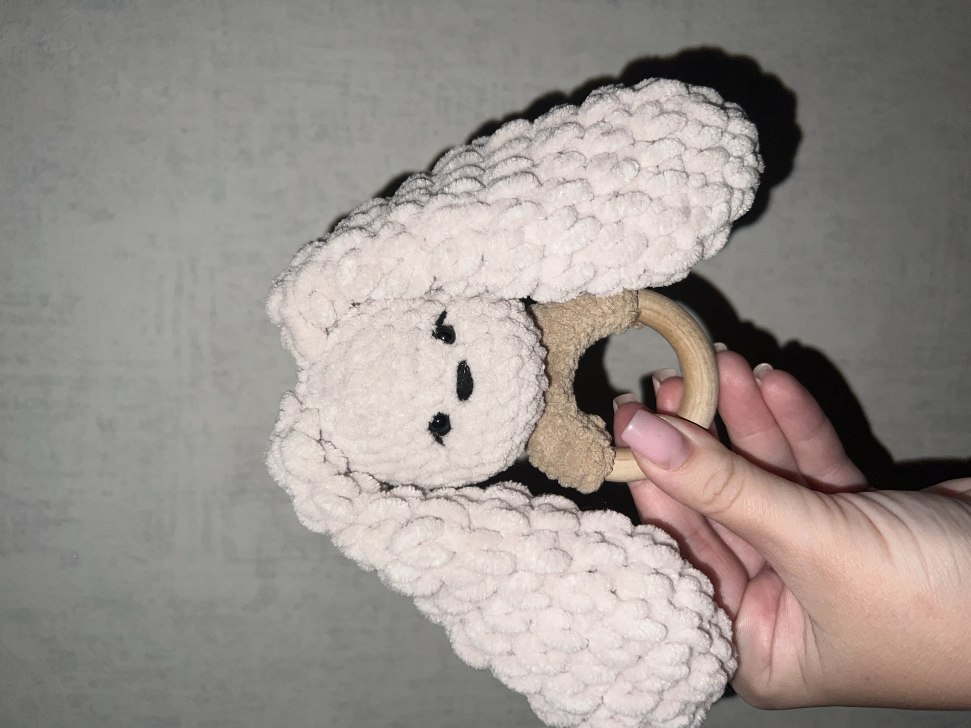 A close-up, vertical, eye-level shot shows a person's hand holding a crochet bunny rattle against a gray, textured background. The bunny, made of light pink, fuzzy yarn, has two long ears that droop down on either side of its head. The bunny's face has two small black eyes and a black stitched nose. A light brown crocheted piece is attached to a wooden ring, which is held in place by the bunny's head. The person's hand, with neatly manicured nails painted a light pink, is positioned below the bunny, gently supporting the wooden ring. The overall lighting is dim, creating a shadow on the wall behind the rattle.