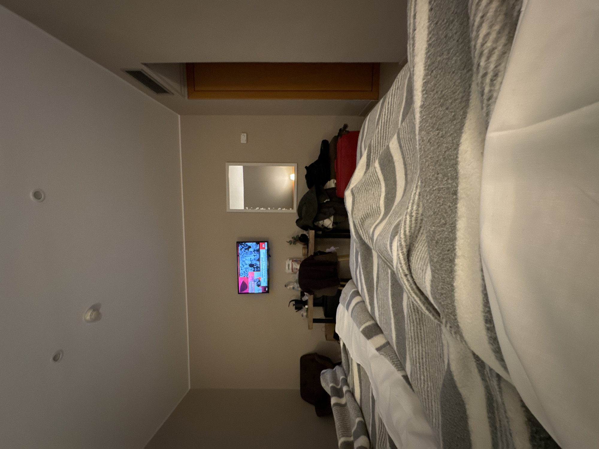 A wide shot of a room with a bed taking up a large portion of the right side. The bed has a striped gray and white blanket, and white sheets. In the background, there is a television on the wall, displaying a news program with red and blue colors. There is a shelf with various items, including clothing, bags, and bottles. The overall lighting is dim, but the television screen provides some illumination. The location of the room in Barcelona, Spain, cannot be determined from this image.