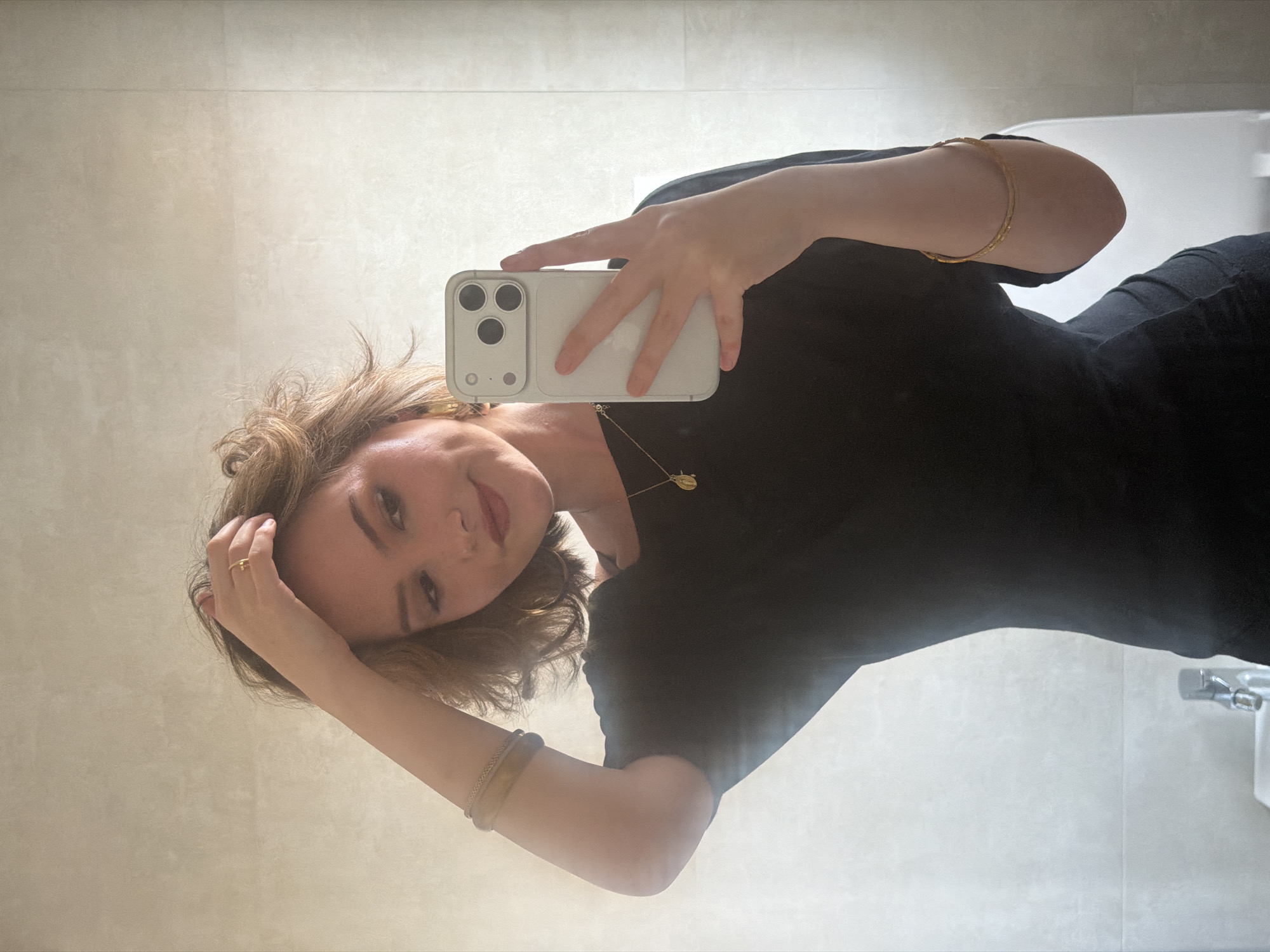 A woman is taking a selfie in a bathroom. She is holding a white smartphone with three camera lenses. She is wearing a black dress and gold jewelry including a necklace, ring, and bracelets. Her hair is blond and styled in waves. The bathroom is tiled in a light color, and a toilet and faucet are visible. The location in the city of Milan, Italy cannot be determined from the provided image.
