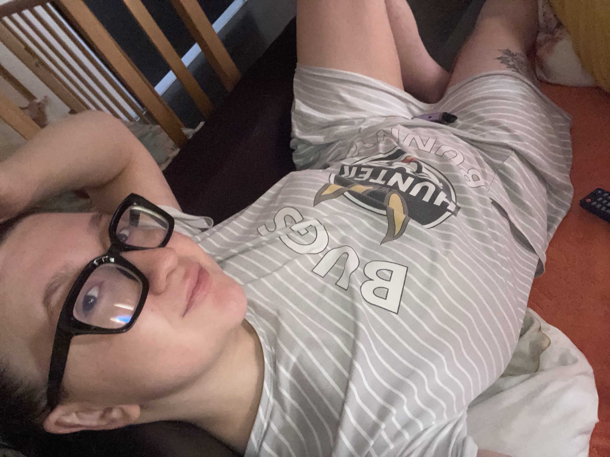 A person wearing black-rimmed glasses and a striped t-shirt with "Bugs Hunter" printed on it is lying down. A remote control is visible on an orange surface next to the person. A crib is in the background. The location cannot be determined based on the image.
