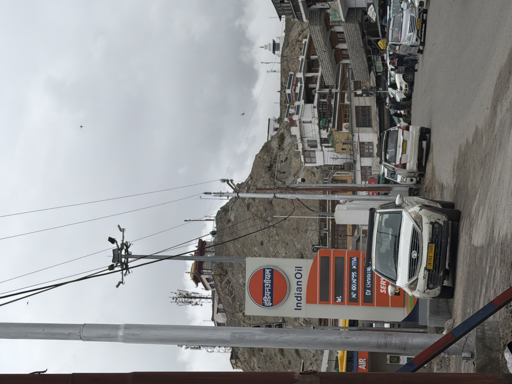 This is a street-level, eye-level, medium shot of a street in Leh, India. The image features a white car with a yellow license plate and an Indian Oil gas station sign. The gas station sign has the Indian Oil logo and the words "Indian Oil" written in English and Hindi. Behind the gas station, there are buildings on a hillside and a cloudy sky. In the background, there are several other vehicles and people standing on the side of the road. The overall scene suggests a town or city setting with signs of commerce and everyday life.