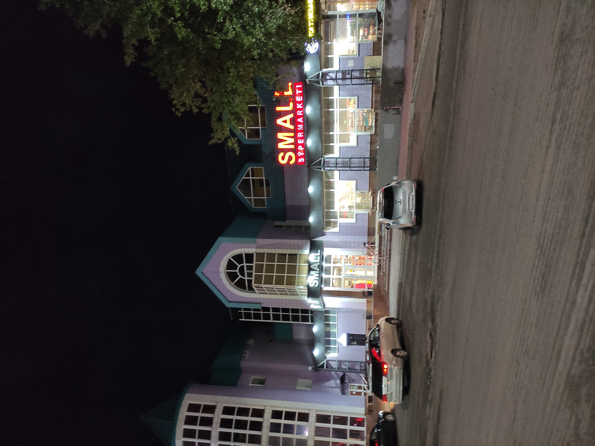 Here's a description of the image:
The photograph captures an evening shot of a building identified as "SMALL Supermarketі" (likely "Small Supermarket" in Russian). This building is located in the city of Pavlodar, Kazakhstan.
The structure itself is a multi-storied building, featuring a combination of modern and somewhat dated architectural styles, painted in lavender with teal accents. Prominently displayed on the building’s facade are the lit-up words "SMALL" in bold red letters.
The street in front of the supermarket is visible, with several parked cars. The lighting suggests a night scene, with streetlights and the supermarket's own lighting illuminating the area. Some greenery (likely trees) is seen overhanging the building. The road is paved and appears to be made of concrete or asphalt.