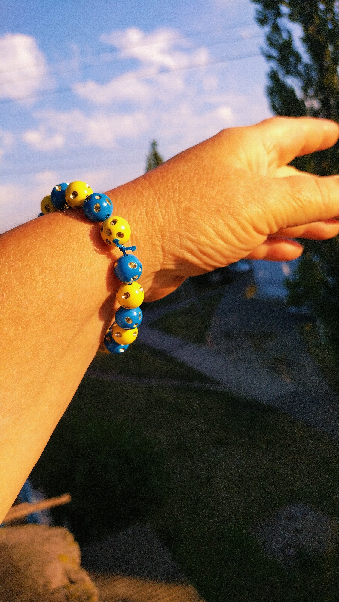 The image shows a close-up of a person's arm and hand wearing a blue and yellow beaded bracelet. The beads are round and appear to have small holes or designs on them. The bracelet is made of alternating blue and yellow beads, suggesting the colors of the Ukrainian flag. The person's skin is illuminated by warm, golden light, indicating it might be sunset or sunrise. The background is out of focus but shows a blue sky with some white clouds, and hints of green foliage and possibly buildings, consistent with an outdoor urban or semi-urban environment. While the specific location within Horishni Plavni, Ukraine is not explicitly identifiable from the visual cues, the overall setting suggests a scene captured in that region.