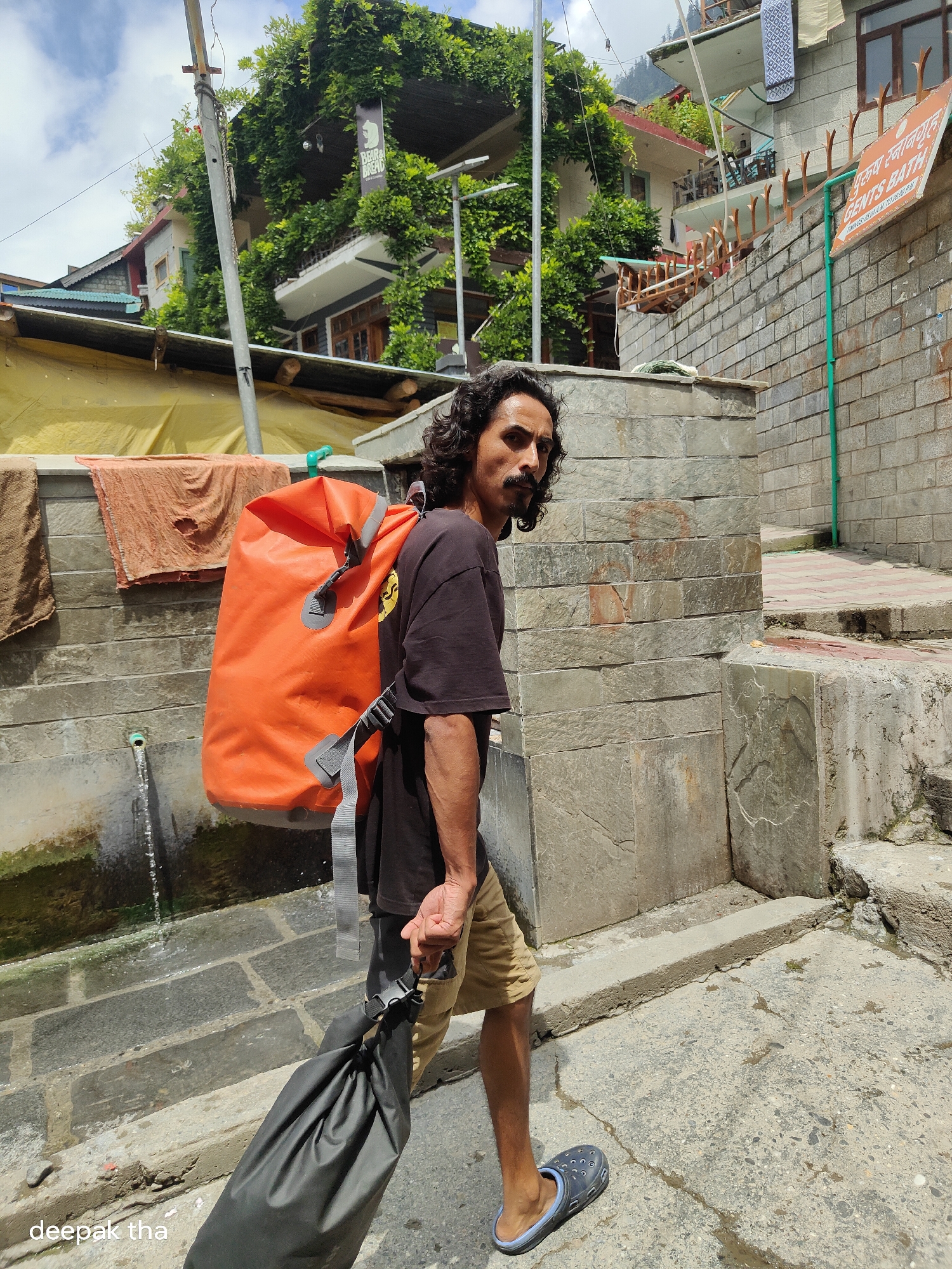 A man with dark, curly hair and a mustache stands on a stone pathway in Vashisht, India, carrying a large orange dry bag on his back and a black dry bag in his right hand. He is wearing a dark t-shirt and tan shorts. Behind him, a building is covered in lush green vines, with balconies and windows visible. A sign on the building reads "GENTS BATH". The environment appears to be outdoors with stone walls and steps, and a slight slope to the ground. Water trickles down a stone channel on the left side of the image. The sky above is partly cloudy.