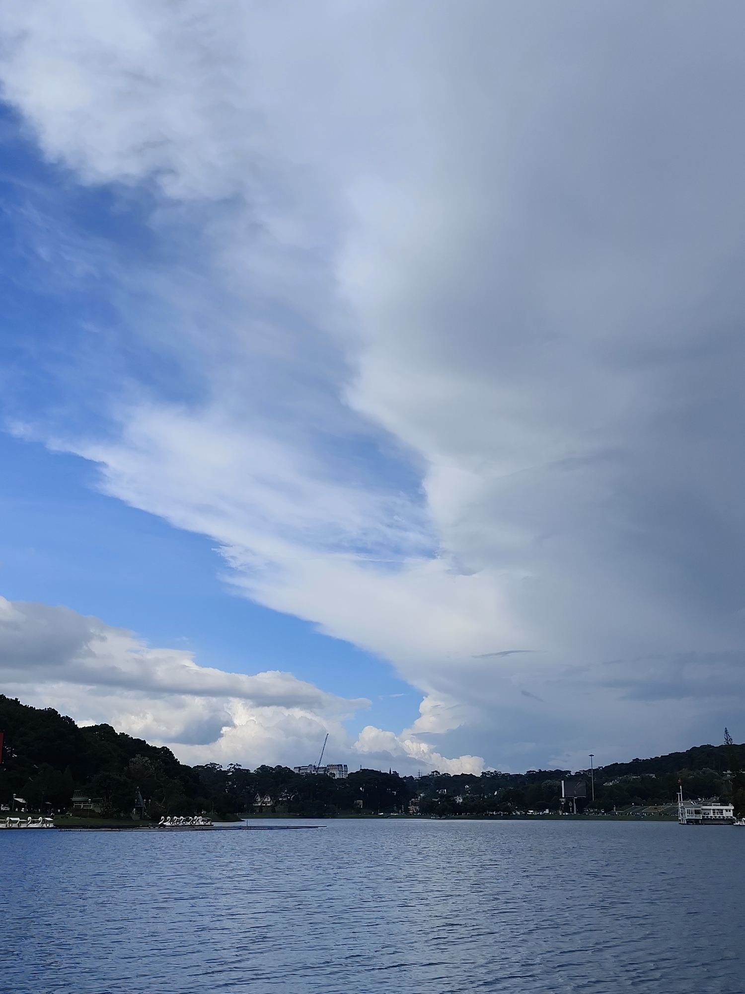 The image depicts Xuan Huong Lake in Da Lat, Vietnam. The foreground is dominated by the calm, blue water of the lake, with gentle ripples visible on its surface. In the mid-ground, the distant shoreline is lined with lush green trees, obscuring much of the town. A few buildings are visible peeking through the foliage, and a tall construction crane stands prominently against the sky on the right side of the frame. The background is a dramatic sky filled with a mix of bright blue patches and heavy, grey clouds, suggesting a change in weather. Several white swan-shaped paddle boats are docked along the left side of the lake, and a white building with a red-tiled roof is situated on the far right shoreline.