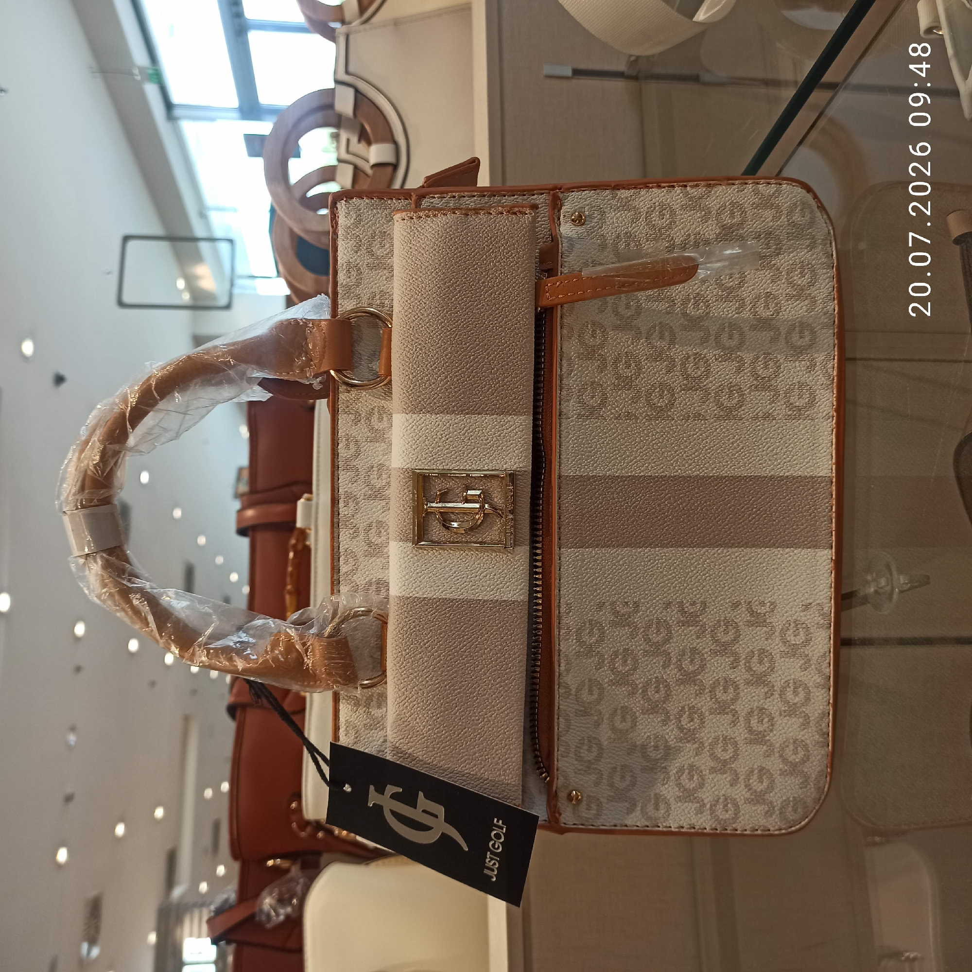 This is a close-up, eye-level, indoor, slightly off-center, full-shot photograph of a handbag. The handbag is a medium-sized, structured tote bag with two top handles, made of a beige material with a repeating pattern of "JG" logos in a lighter beige. It features a central vertical stripe of darker beige. The handbag has a zip closure across the top of the main compartment, and a flap pocket on the front. A black tag with a silver "JG" logo and the words "JUST GOLF" is attached to the handle. The handbag is placed on a glass surface, likely a display shelf in a retail store. In the background, other handbags and store fixtures are visible, along with fluorescent lighting on the ceiling and a large window. The timestamp "20.07.2026 09:48" is visible in the bottom right corner of the image. The location is identified as Bari, Italy.