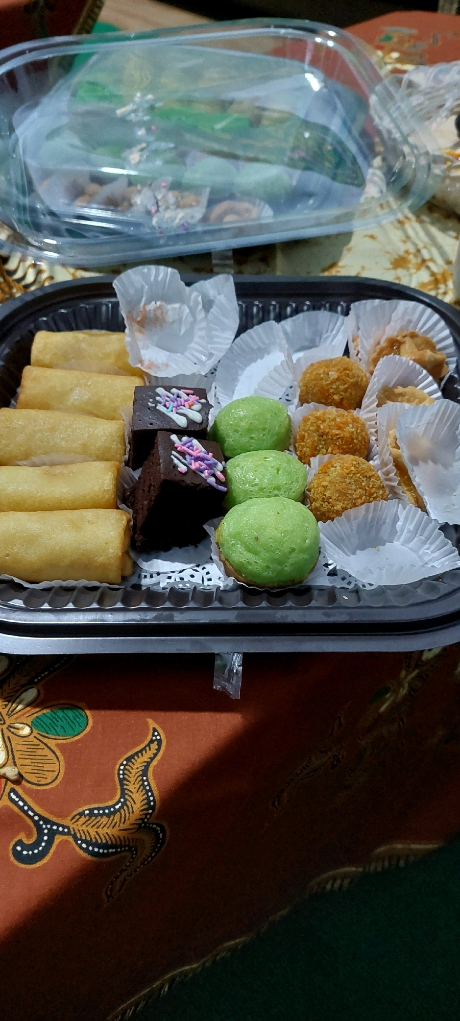 A close-up, high-angle shot captures a black plastic tray filled with various snacks. The tray is positioned on a patterned surface, possibly a tablecloth.
The snacks are arranged in rows and compartments. On the left side, there are several golden-brown, cylindrical spring rolls. Next to them are small, dark brown square brownies topped with white and pastel-colored sprinkles. To the right of the brownies are round, green cake-like snacks. Further to the right are several bite-sized, golden-brown, breaded snacks, resembling croquettes or fried dumplings. In the far right of the tray, there are some small tarts with a golden filling and a crimped crust.
The snacks are mostly placed in white paper liners. A clear plastic lid, slightly askew, covers a portion of the tray in the background. The patterned surface beneath the tray features floral and abstract motifs in shades of orange, brown, and green. The overall lighting suggests an indoor setting. There are no people or animals visible in the image. The exact location is unknown.