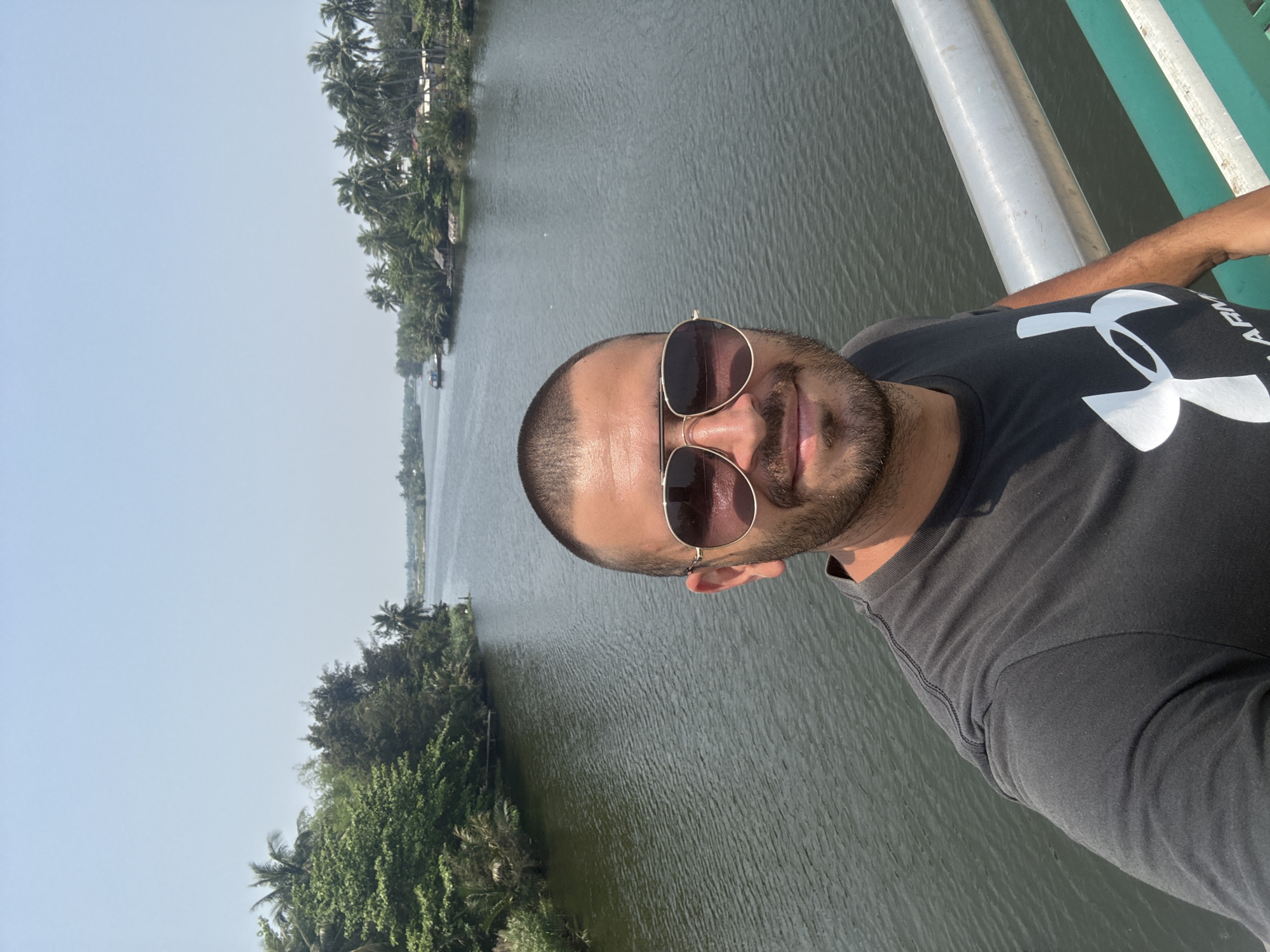 A man wearing sunglasses and a black Under Armour T-shirt is taking a selfie on a bridge. The body of water and lush vegetation on either side of the bridge suggest he might be near the Cổ Cò River, which is located between Da Nang and Hoi An in Vietnam.