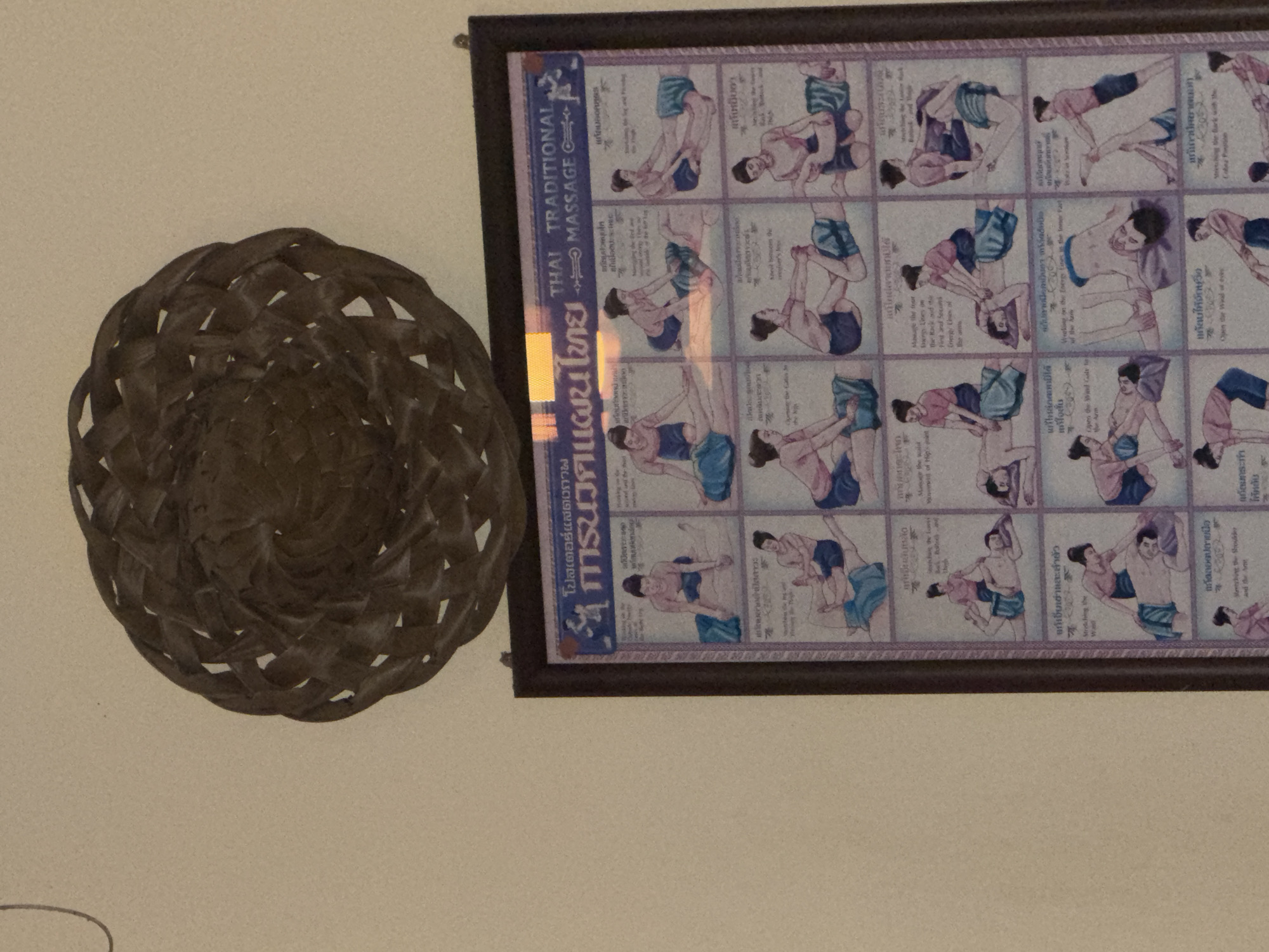The image shows a framed poster depicting the techniques of traditional Thai massage. The poster is hung on a wall, and above it, a woven decorative item made of natural materials is suspended. The poster itself is divided into a grid of illustrations, each showing two figures engaged in a specific massage posture or action. The illustrations are rendered in a watercolor style, with figures depicted in blue and pink attire. The poster also features text in both Thai and English. The overall setting appears to be indoors, possibly a spa or massage establishment in Baan Tai, Thailand. The lighting suggests ambient indoor lighting.