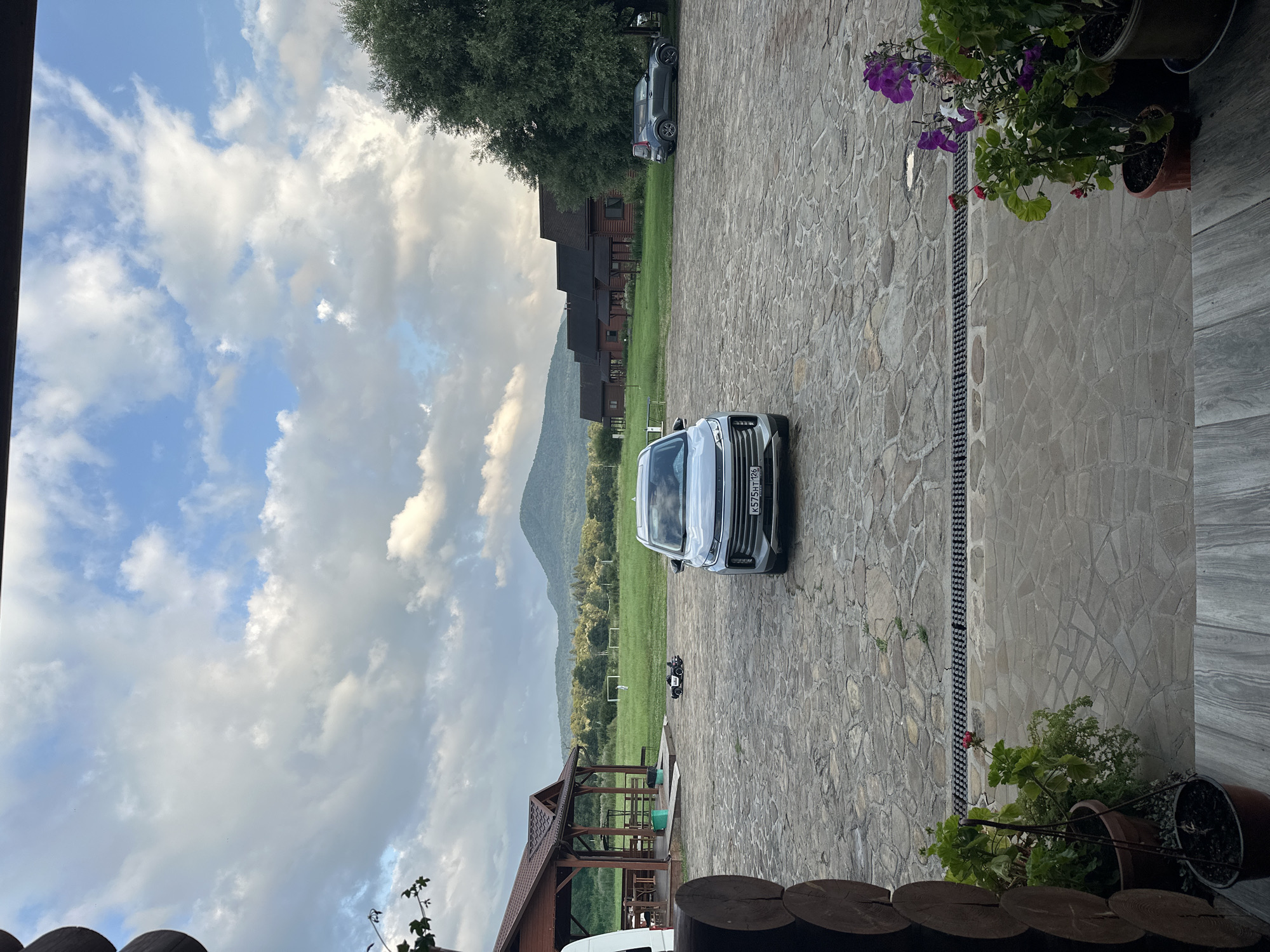 A modern, silver SUV is parked on a stone-paved courtyard in front of a rural establishment. The SUV's license plate, K575HT 126, is visible. To the left of the SUV, a small, black toy police car is placed on the ground. In the background, a covered wooden structure with tables and chairs is situated. Further back, a row of wooden houses with dark roofs stretches across a green field. In the distance, a mountain range with lush green slopes under a cloudy sky dominates the horizon. The scene appears to be set in the city of Dakhovskaya, Russia. Potted plants with purple and red flowers are visible in the foreground.