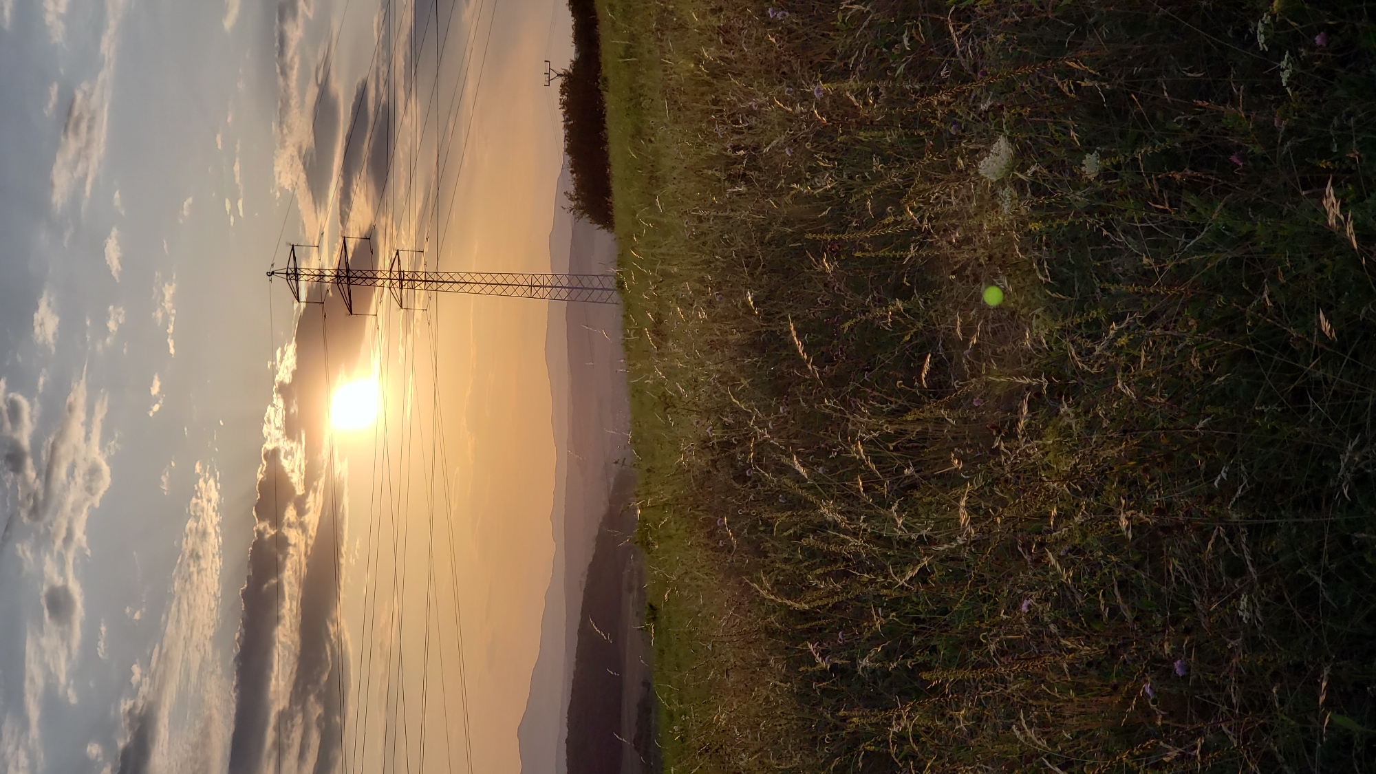 The image captures a sunset scene with a tall electricity pylon in the foreground, silhouetted against a sky filled with clouds and the setting sun. The sun is positioned towards the left side of the frame, casting a warm, golden light across the landscape. A field of tall grass and wildflowers occupies the lower portion of the image, with some blades of grass illuminated by the sunlight. In the distance, rolling hills and mountains are visible, layered in soft hues of orange and purple due to the atmospheric conditions of sunset. The pylon is a prominent feature, with power lines stretching horizontally across the frame. The overall impression is one of a rural landscape at dusk, with industrial elements integrated into the natural scenery. This image is a photograph of a landscape in the vicinity of Prešov, Slovakia.