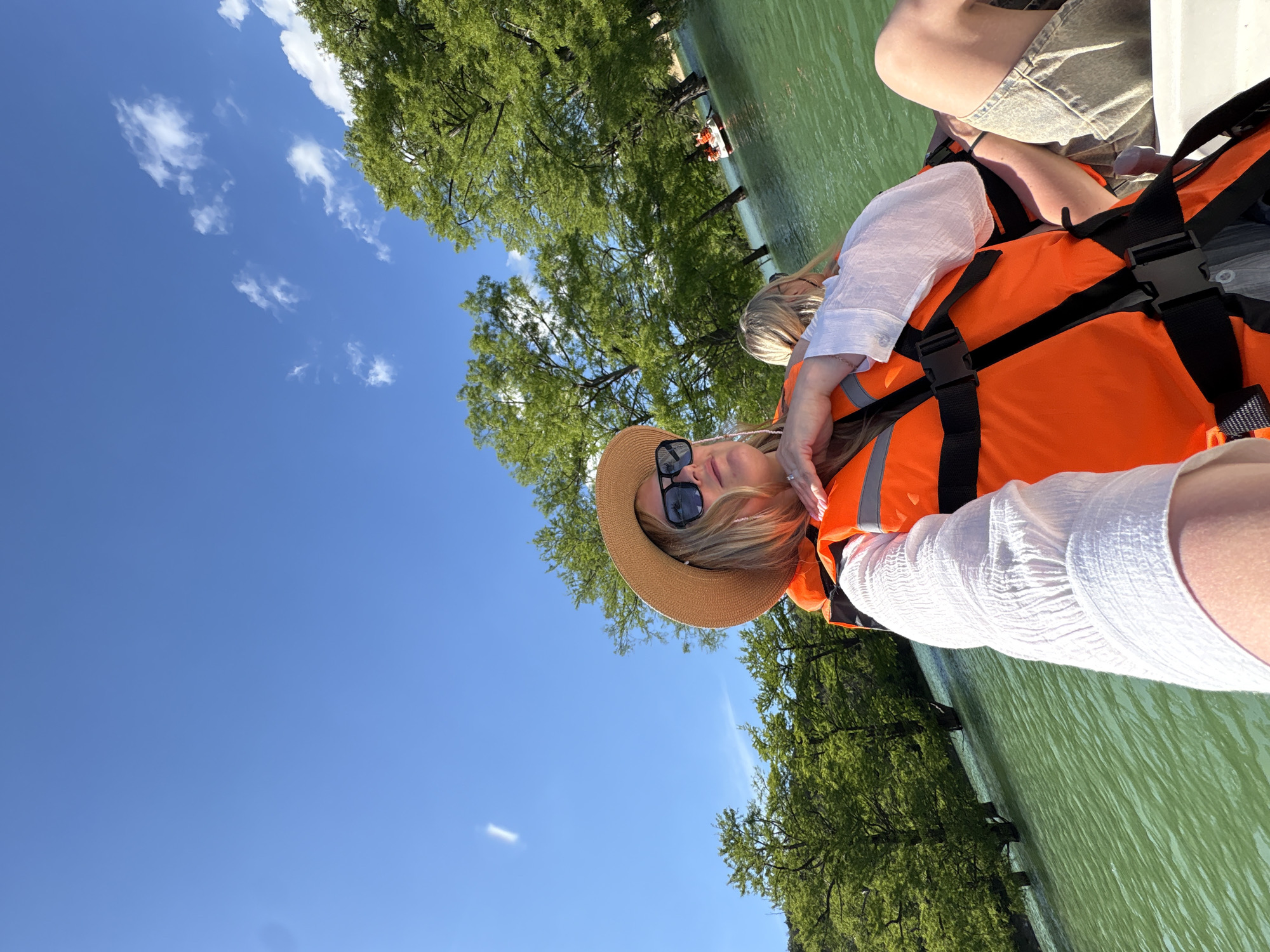 This media file captures a bright and sunny outdoor scene on a body of water, likely the Cypress Lake in Sukko, Russia.
The main subject is a woman positioned in the foreground, looking towards the camera. She has blonde hair and is wearing a wide-brimmed straw hat, dark sunglasses, a textured white long-sleeved shirt, and a bright orange life vest securely fastened. A white ring is visible on her left ring finger. Behind her, another person is partially visible, also wearing an orange life vest, suggesting they are on the same white boat or watercraft.
The water itself is a calm, greenish color, reflecting the surrounding greenery. The banks of the waterway are lined with numerous lush green trees, possibly cypress, consistent with the provided location context. Further in the distance on the water, a smaller white boat carrying at least two more individuals, also equipped with orange life vests, can be seen.
The sky is a clear, vibrant blue with scattered, wispy white clouds, indicating fair weather. The bright sunlight suggests the time of day is likely midday or early afternoon, ideal for outdoor water activities. The overall mood appears relaxed and enjoyable. There is no visible text in the image.