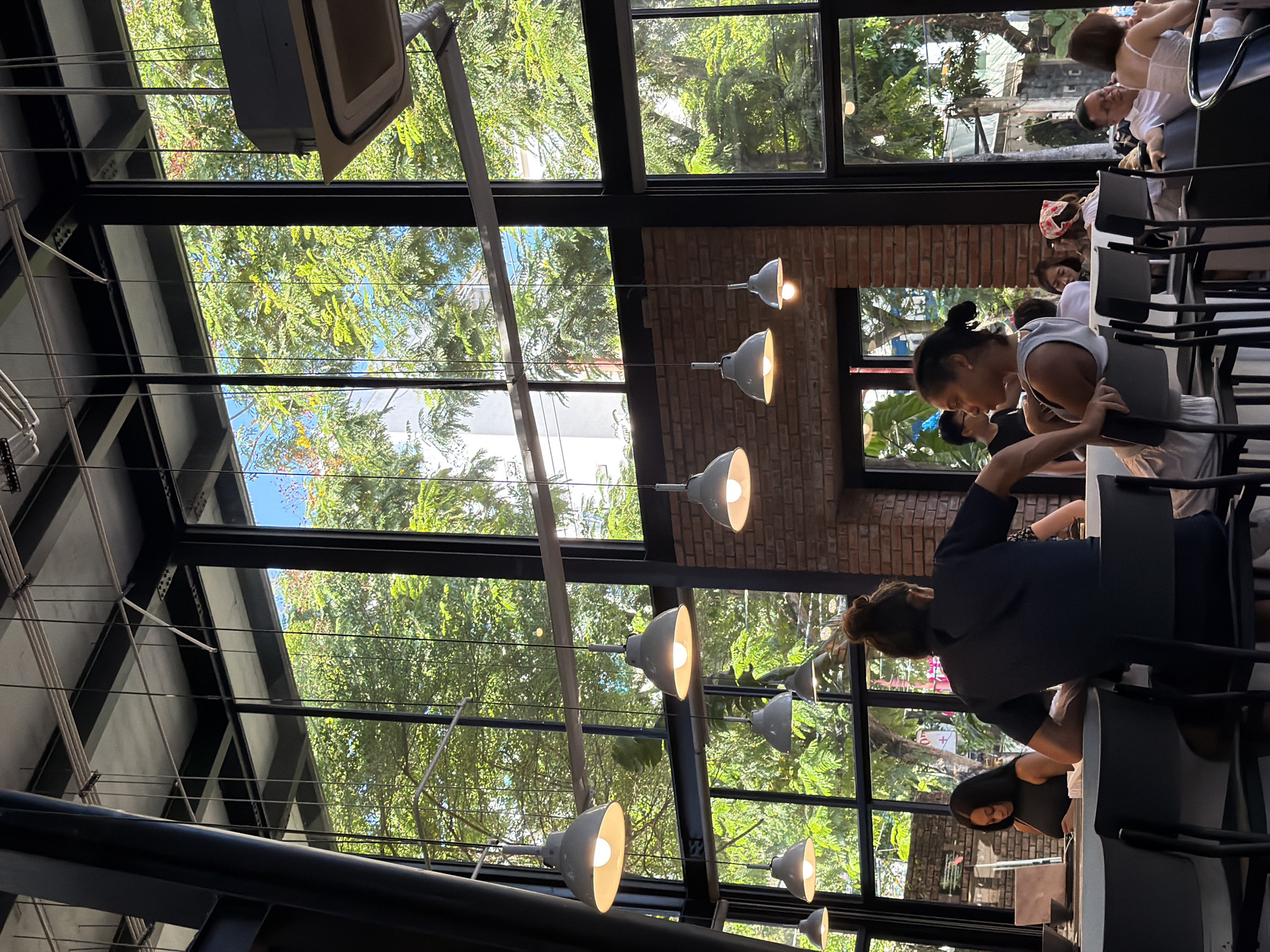 The photograph captures the interior of a café in Đà Nẵng, Vietnam, characterized by its abundance of natural light filtering through large glass windows framed by black metal. Lush greenery and trees are visible outside, suggesting an urban setting with a connection to nature.
Several people are present, engaged in activities such as sitting at tables, conversing, and seemingly enjoying the café's ambiance. The interior features exposed brick walls and a series of hanging pendant lights, which contribute to the café's aesthetic.
The café appears to be bustling, indicating its popularity. The scene conveys a sense of relaxation and socializing.