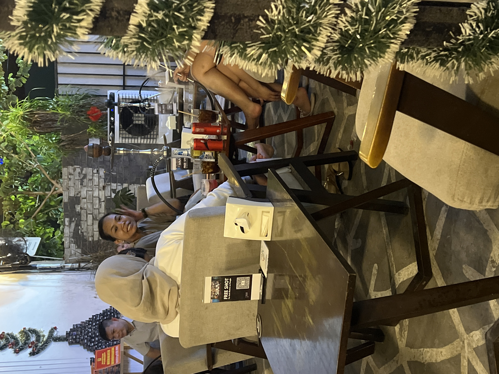 The image shows an outdoor cafe scene with multiple people. A woman in a hijab sits at a table with a man, while another woman sits nearby and looks toward the camera. The table has marketing materials including a "FREE SHOT" promotion. Another table has legs dangling over it, suggesting elevated seating. In the background, a decorative Christmas-themed arrangement is visible. A fan provides airflow. Based on this analysis, the image captures a casual social scene in a public space. No information is presented in the prompt that would verify the location in the city of Đà Nẵng, Vietnam.