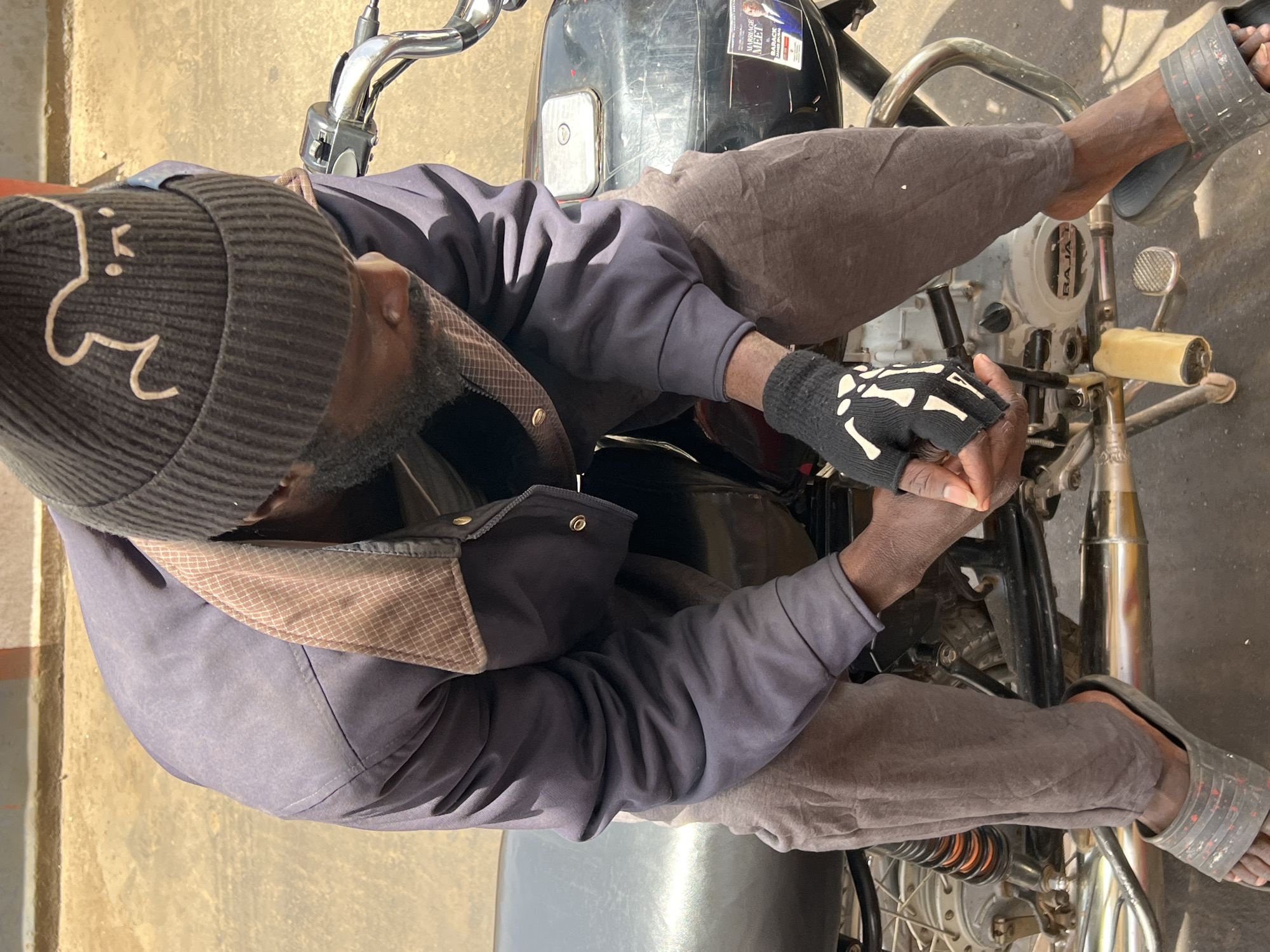 A man is sitting on a black and silver motorcycle. He is wearing a dark gray beanie with a white design on it, a blue jacket, gray pants, and sandals. One of his hands is covered by a black glove with a white skeleton hand design. The motorcycle has a sticker for "Marriage Meet Barak Obama". The location is Jos, Nigeria.