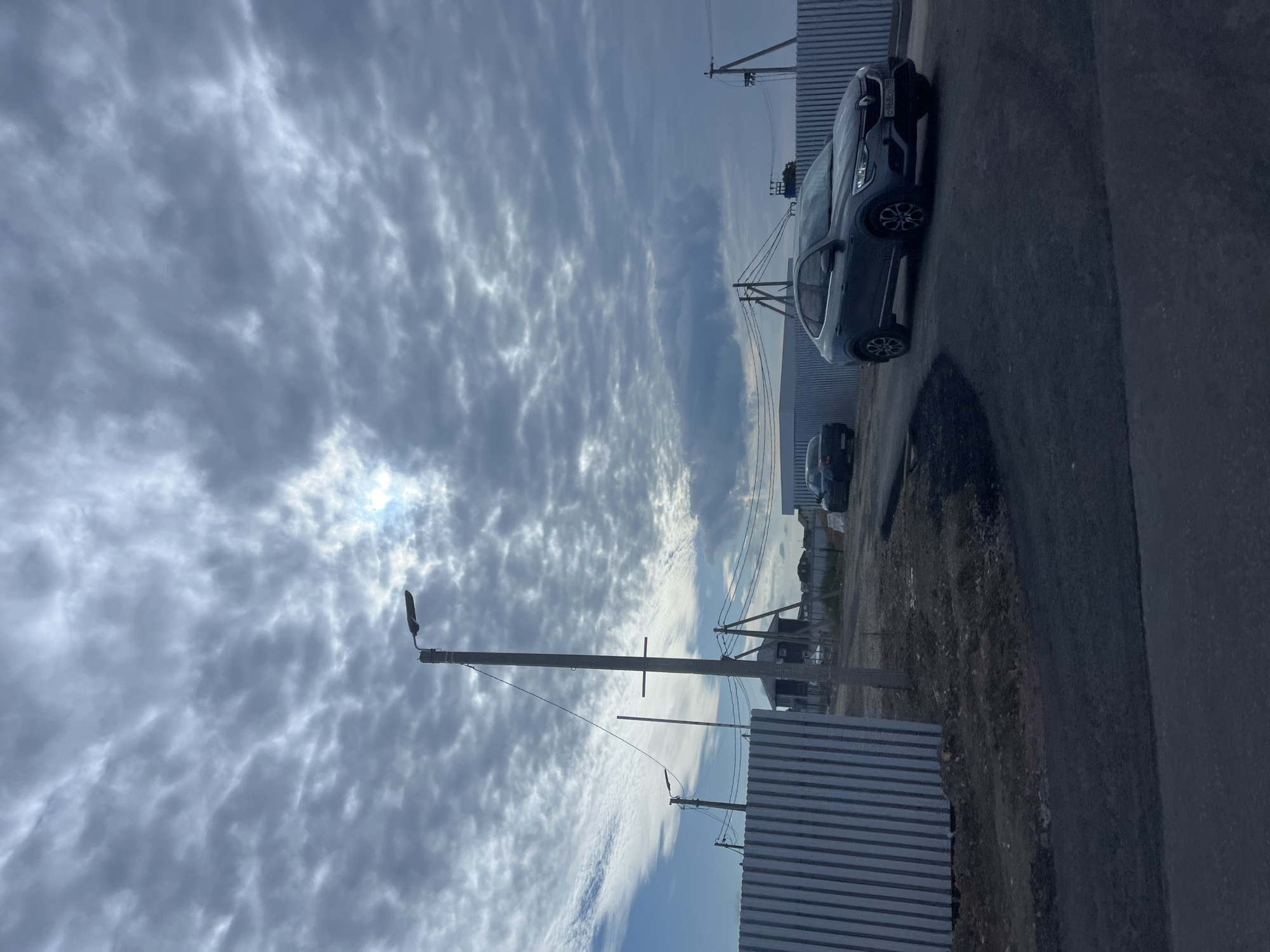 The image depicts a construction site in Glyadino, Russia, under a cloudy sky. In the foreground, a dark grey car is driving on an asphalt road. Beyond the road, a metal fence runs horizontally across the frame. Further back, another car is parked near a house. A tall lamppost stands to the left of the frame. In the background, power lines and more houses are visible, indicating a developing residential area. The sky is filled with textured clouds, with a small patch of bright light suggesting the sun is attempting to break through.
