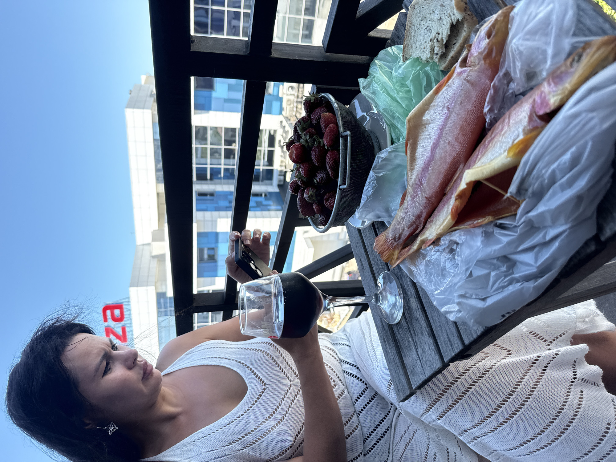 A woman with dark hair, wearing a white crocheted dress, sits at a wooden table on a balcony. She is looking down at her phone. On the table, there is a glass of red wine, a metal bowl filled with strawberries, two raw fish wrapped in plastic, and a slice of bread. In the background, a modern building with blue windows and red lettering that reads "IZZA" is visible against a clear blue sky. The railing of the balcony is dark wood. The scene appears to be set in Sochi, Russia, given the context of the building signage.