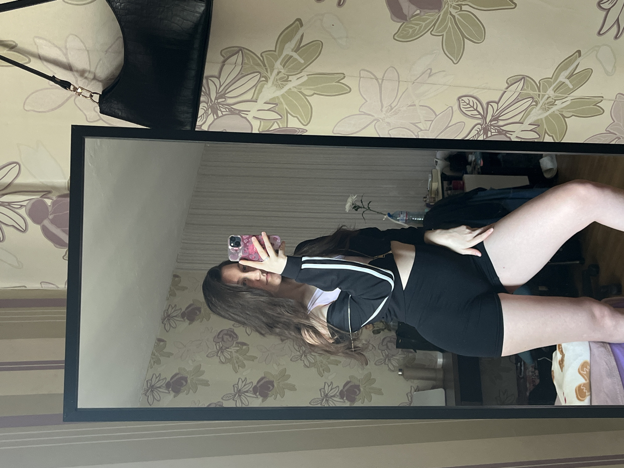 A mirrored reflection shows a young woman indoors in an Unknown, Unknown location. She holds a cell phone up to take a selfie, wearing a black athletic suit with white stripes down the sleeves of her jacket. Her hair is long and brown. The background behind the mirror shows a floral wallpaper. A black purse hangs in the top left corner. The rest of the room is visible through the mirror and shows bedding and furniture. A water bottle and flower are also visible behind her.