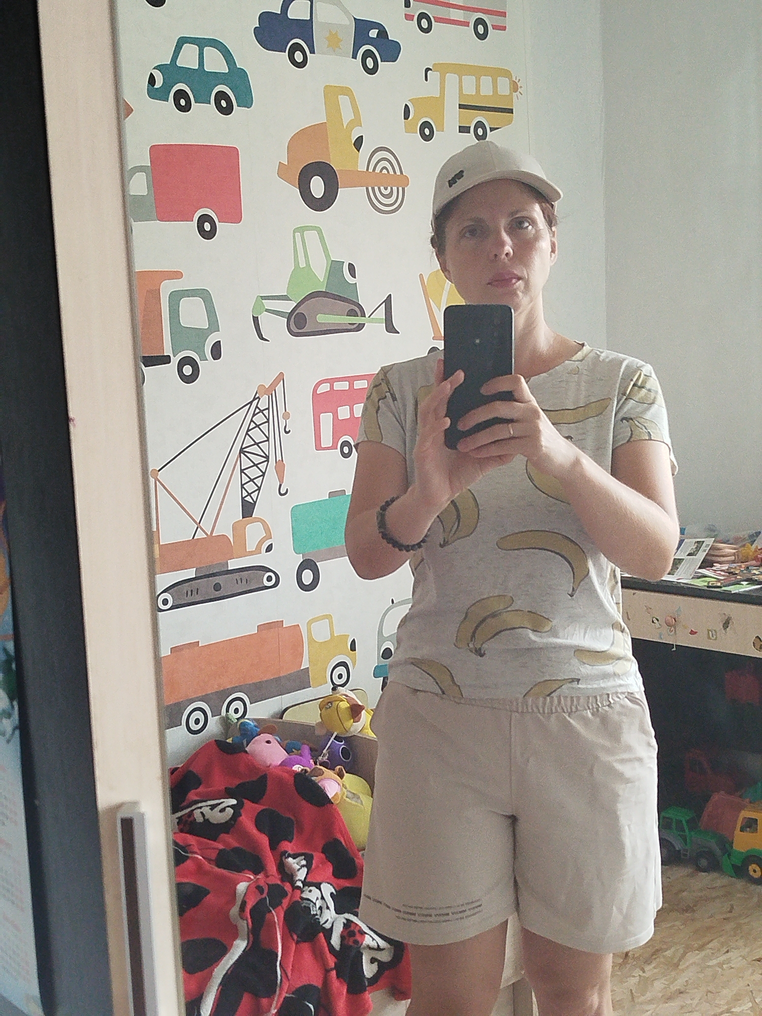 A woman wearing a light-colored baseball cap and a t-shirt with a banana print takes a selfie in a room with children's wallpaper depicting various vehicles. She is holding a black smartphone in her right hand. In the background, to her left, is a closet with a mirror, reflecting a portion of the room and her. The wallpaper features a variety of trucks, cars, and construction vehicles in bright colors. To the right of the woman, a shelf is visible with some toys and books. A red blanket with black spots is draped over a surface in the lower left portion of the image. The flooring appears to be light-colored wood. The woman's lower body is visible, showing she is wearing light-colored shorts. The overall environment suggests a child's room or play area. The location is the city of Zavodoukovsk, Russia.