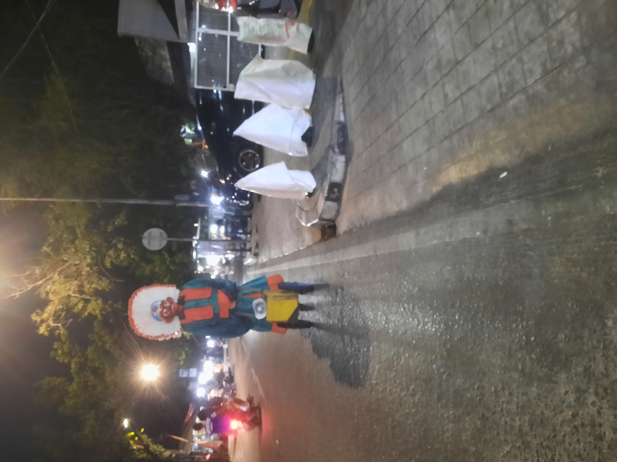 At night on a street in North Jakarta, Indonesia, a person in a colorful costume with a large headdress stands near the center of the frame. The costume consists of a blue garment with orange accents and a yellow cart or box attached to the front. The person is standing on the asphalt road, which appears wet. To the right of the street, a sidewalk is visible with several white, sheet-covered objects placed on it. In the background, trees and streetlights are illuminated, and several motorcycles and cars are present, suggesting a busy urban environment. The overall scene is dimly lit, with artificial lights providing illumination.