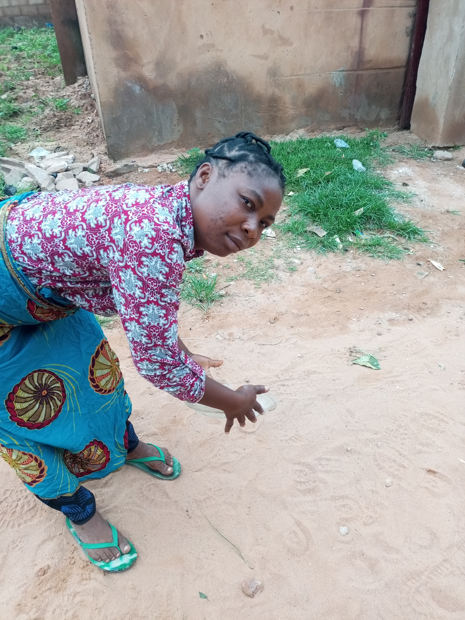 A young Black woman, with her hair in braids, wearing a patterned red and white long-sleeved shirt and a blue skirt with a circular pattern, is in a crouched position. She is holding a clear plastic bottle in her right hand and appears to be interacting with the sandy ground in front of her. Her feet are clad in green flip-flops. The background consists of a weathered, light-brown wall and some patches of green grass. Debris, including rocks and scattered white objects, is visible in the environment. The scene is set outdoors on sandy terrain, consistent with a location in Tempure, Nigeria.