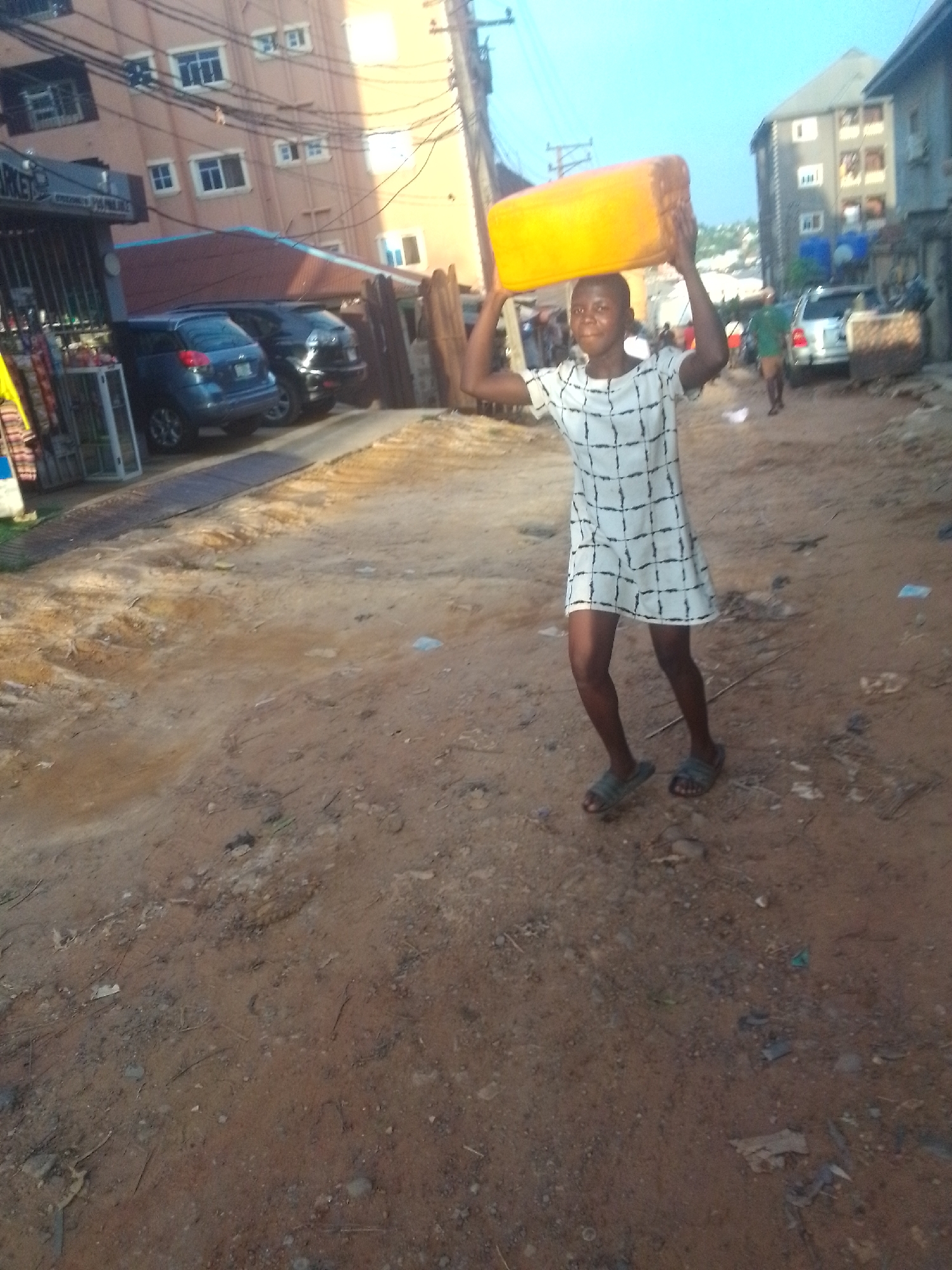 A young person in a white dress with a black grid pattern is carrying a yellow rectangular container on their head, walking on a dirt road in Nkpor, Nigeria. They are wearing sandals. The road is dusty and uneven. In the background, there are buildings, cars, and a few other people walking. A market stall is visible on the left side of the frame. There are power lines above.