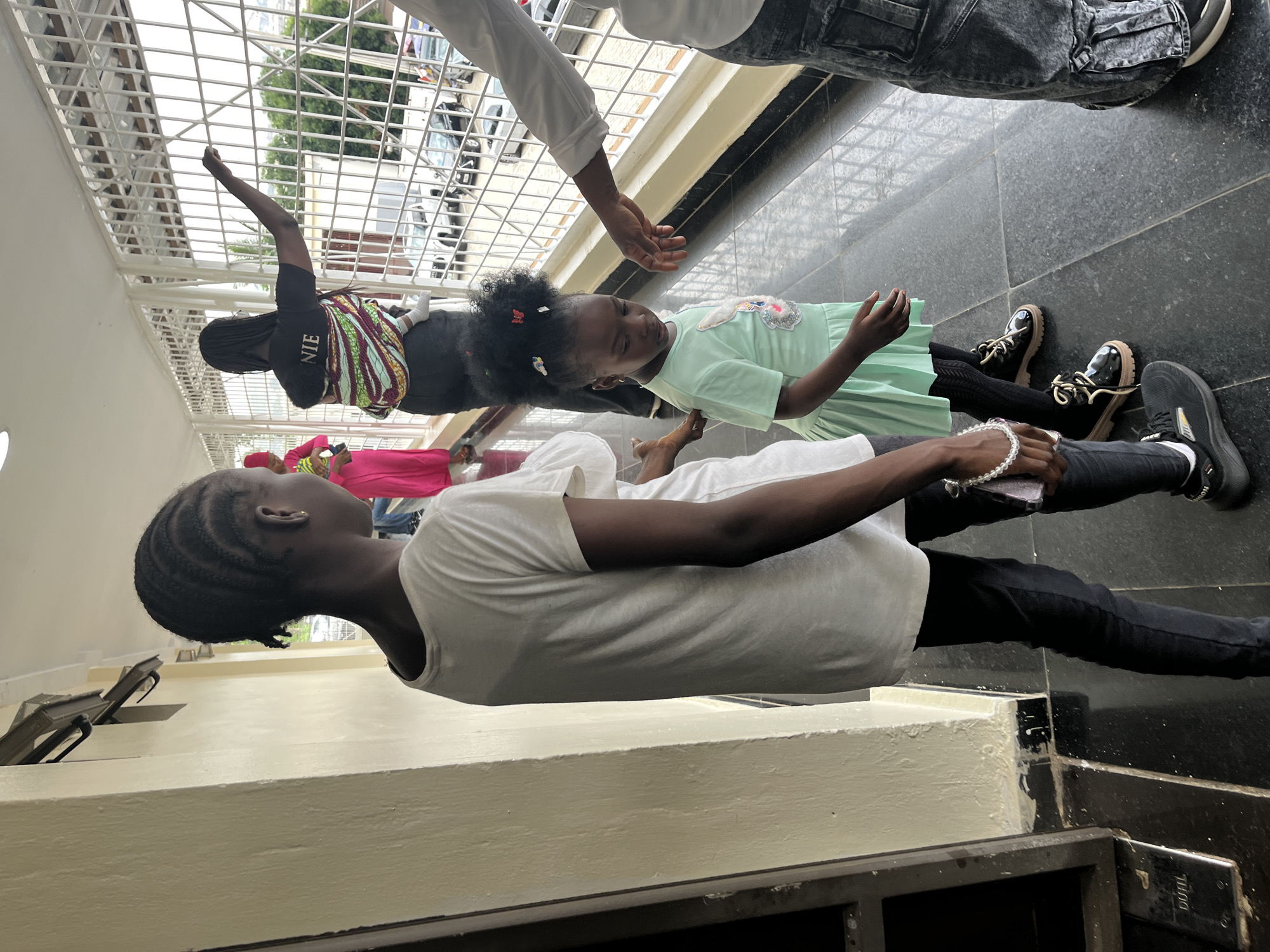 The image depicts an outdoor hallway in what appears to be a modern building, likely in Abuja, Nigeria. A young girl in a light green dress stands in the foreground, looking down. To her left, a girl with braided hair in a white t-shirt and black leggings stands with her back to the camera, holding a beaded bracelet. To the right, the lower half of a person wearing dark, baggy pants and a white shirt is visible, reaching out a hand towards the younger girl. In the background, a woman is seen carrying a baby on her back, wearing a colorful fabric wrap and a black shirt with "NIE" printed on it. She is looking out through a white metal grate that encloses the hallway, which overlooks a street with cars. The hallway floor is tiled in a dark, reflective material. The building has a cream-colored wall on the left side of the frame.