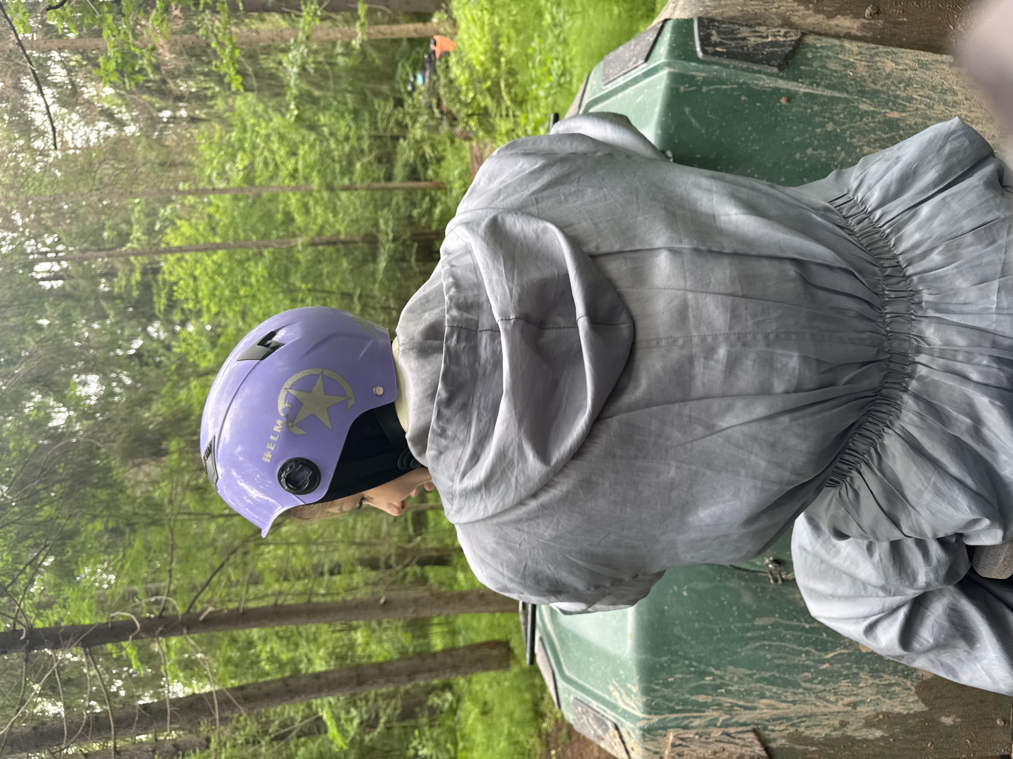 A person, viewed from the back and turned slightly left, is seated on a muddy, dark green all-terrain vehicle (ATV) or utility terrain vehicle (UTV) in a dense forest in the city of Perm, Russia. The individual wears a light purple protective helmet, featuring "HELMET" in white lettering and a light-colored five-pointed star emblem on the left side, above a black adjustment dial. A black balaclava is visible beneath the helmet, covering the lower face and neck. The person is clad in a grey, hooded, full-body protective jumpsuit with elastic gathering at the waist. The vehicle's dark green surface is heavily splattered with dried mud. The environment comprises a lush, vibrant green forest with tall deciduous and coniferous trees, and a dirt track extends into the background. Diffused natural light illuminates the scene.