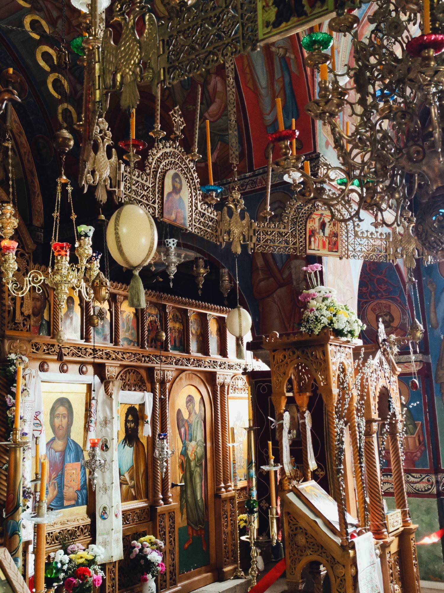 The image shows the interior of an Eastern Orthodox church, likely in Trebinje, Bosnia and Herzegovina, given the architectural style and artistic details characteristic of such churches in the region. The scene is richly decorated with ornate gold-toned chandeliers and hanging lamps.
The focal point is the iconostasis, a screen decorated with religious icons, which separates the nave from the sanctuary. Several icons are visible, depicting religious figures. The wood of the iconostasis is ornately carved.
On the right side of the image is a wooden lectern or pulpit, also elaborately carved, holding a book. Floral arrangements are placed at the base of the iconostasis and on the lectern. The walls are adorned with religious frescoes or murals.
Candles are present throughout the scene, some lit and some unlit, adding to the religious atmosphere. Overall, the image conveys a sense of deep religious tradition and opulence within the church.