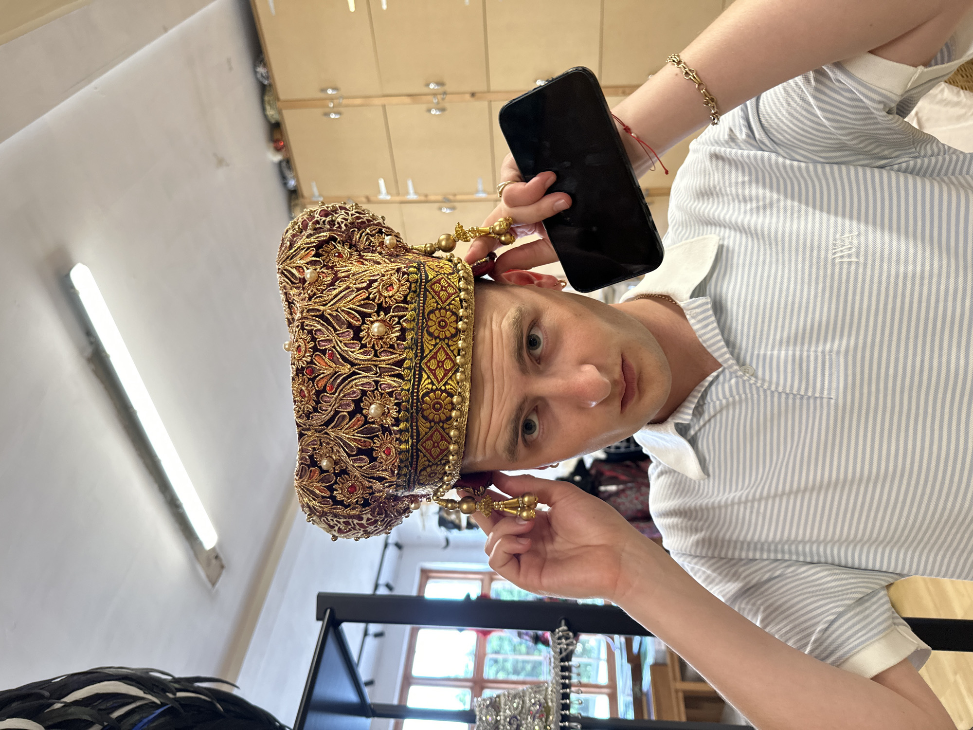 A person is seen wearing an ornate crown-like headdress decorated with gold and red detailing. They are holding a black smartphone in one hand, and the other hand is adjusting a decorative piece of the headdress. The person is wearing a blue and white striped polo shirt. The background includes elements of a retail environment, with display racks of jewelry and other items visible. The location appears to be a store or shop, possibly in the city of London, United Kingdom, given the style of merchandise.