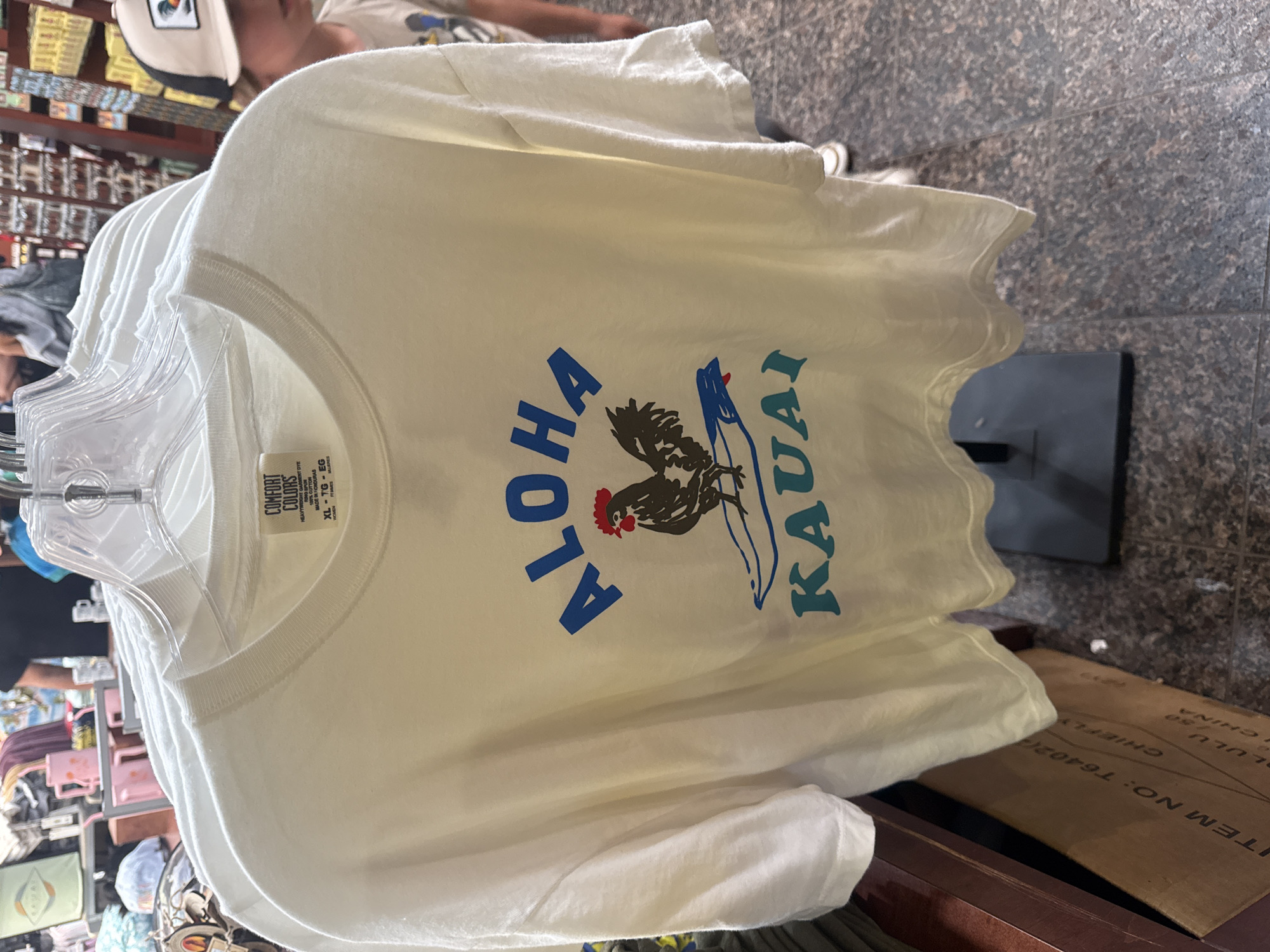 The image displays a white short-sleeved t-shirt hanging on a clear plastic hanger, supported by a black metal display stand, positioned within a retail store in Lihue, Kauai, United States of America. The t-shirt is an XL "Comfort Colors" brand garment, made of 100% cotton in Honduras, and features a central graphic design. This design includes the word "ALOHA" in blue capital letters, a black rooster with a red comb standing on a blue surfboard, and the word "KAUAI" in blue-green capital letters below.
A brown cardboard box, partially visible in the bottom-left foreground, displays text including "ITEM NO: T6402(2)", "CHIEFLY", "LULU SG", and "CHINA". The floor beneath the stand and box is composed of dark gray speckled tiles.
The background reveals typical retail store elements. To the left, shelves contain various merchandise, including folded apparel, light green and pink mugs, and other items. A person wearing a dark shirt is partially visible further back. To the right, shelves are stocked with numerous small items, likely souvenirs or postcards. A young individual, wearing a beige and black trucker hat adorned with a rooster emblem and a light-colored t-shirt, is partially visible on the right side of the frame, facing away from the primary subject.