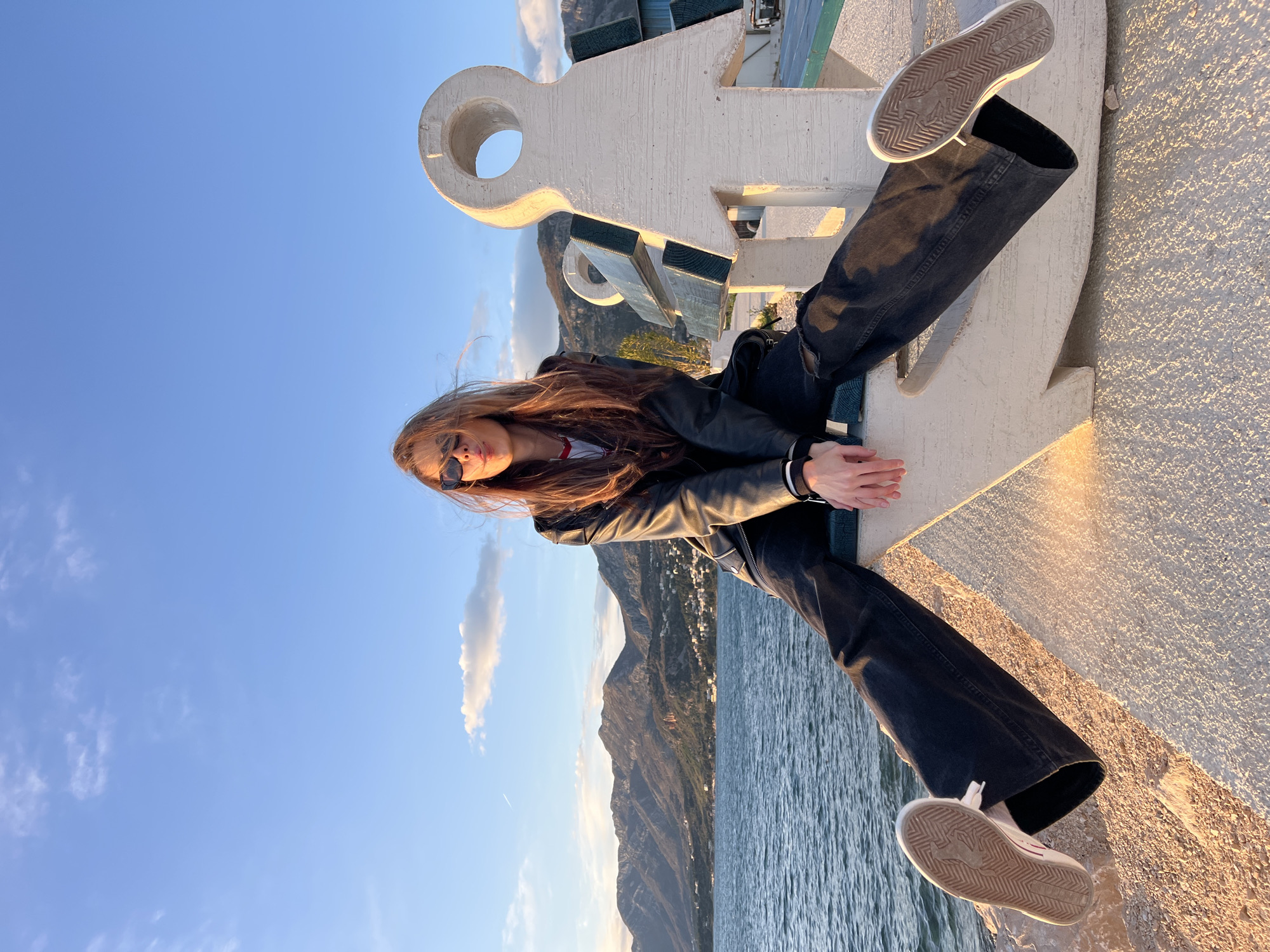 A woman wearing a black leather jacket, black pants, and sneakers is sitting on a large, white, stylized anchor monument. The monument is on a concrete structure, likely a pier or waterfront promenade, in Bar, Montenegro. The woman is looking at the camera and is also wearing sunglasses. The scene includes a view of the Adriatic Sea, mountains, and a clear blue sky with a few clouds. Buildings are visible along the coast. The photograph is taken in the late afternoon or early evening, based on the angle of the sun and warm light.